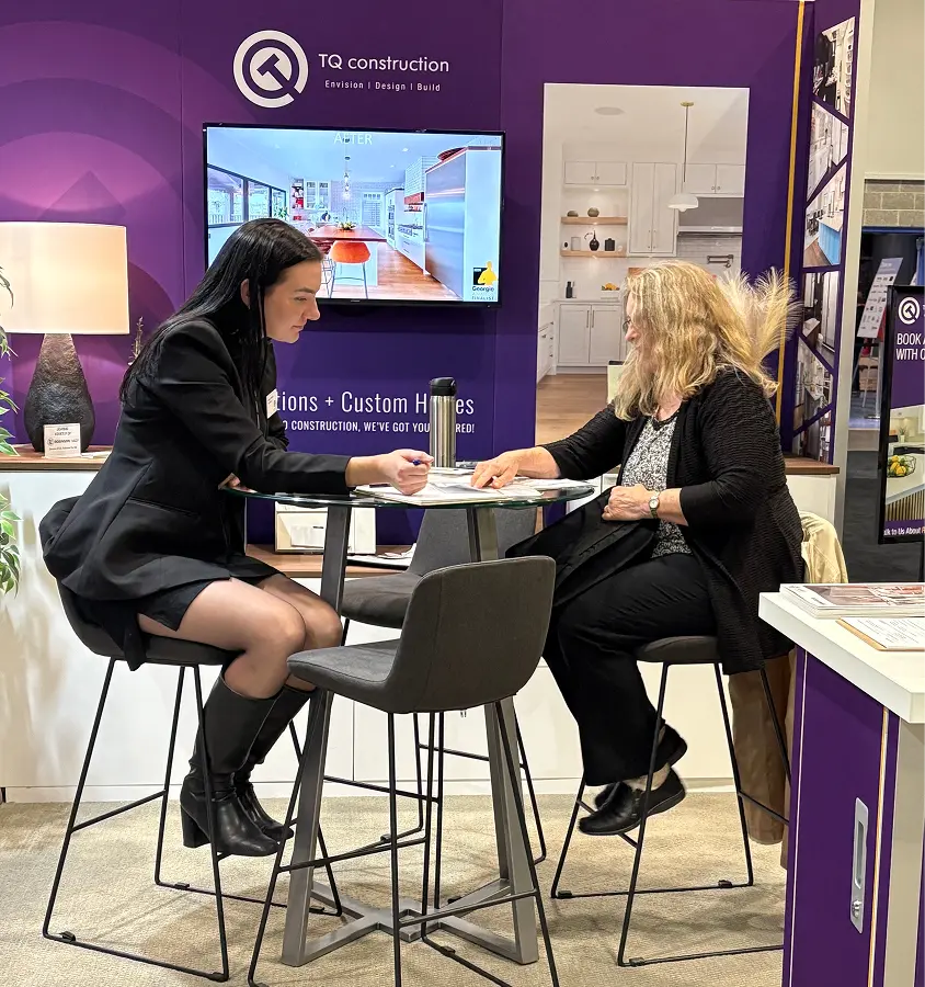 Two women sitting at a round glass table discussing paperwork at a TQ Construction booth with a kitchen renovation display on a purple background.