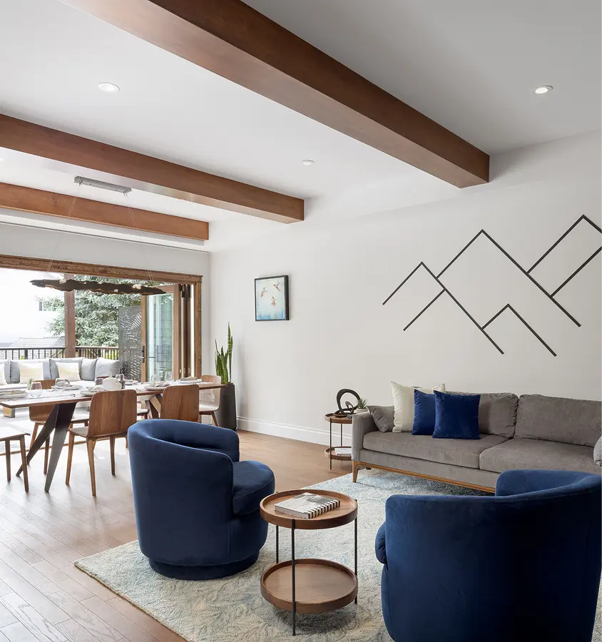 Modern living room with gray sofa, blue armchairs, wooden ceiling beams, and a dining area near sliding glass doors.