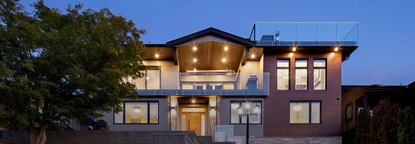 Modern two-story house with large windows and glass balcony railings illuminated at dusk.