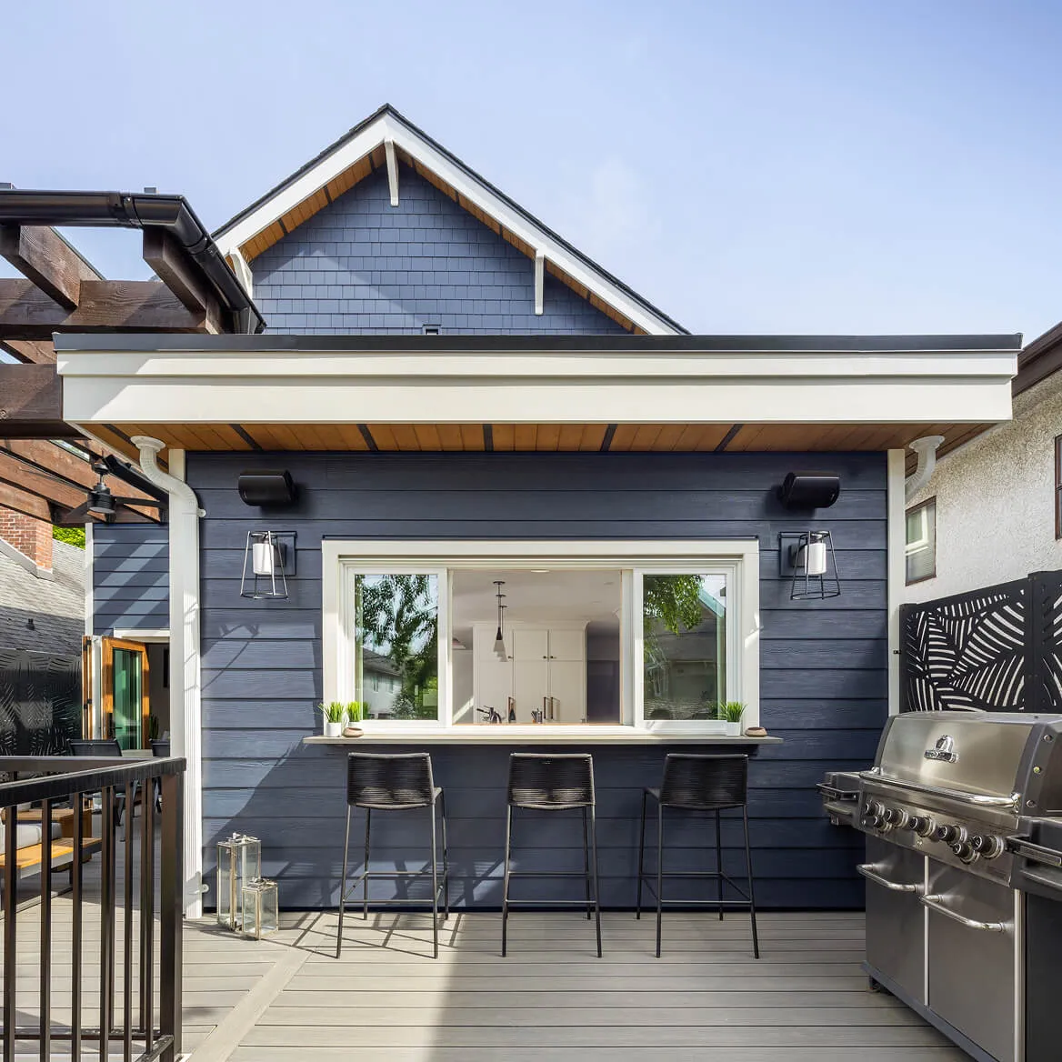 Outdoor patio bar with three black stools, a window serving counter, and a large stainless steel grill next to a blue house.
