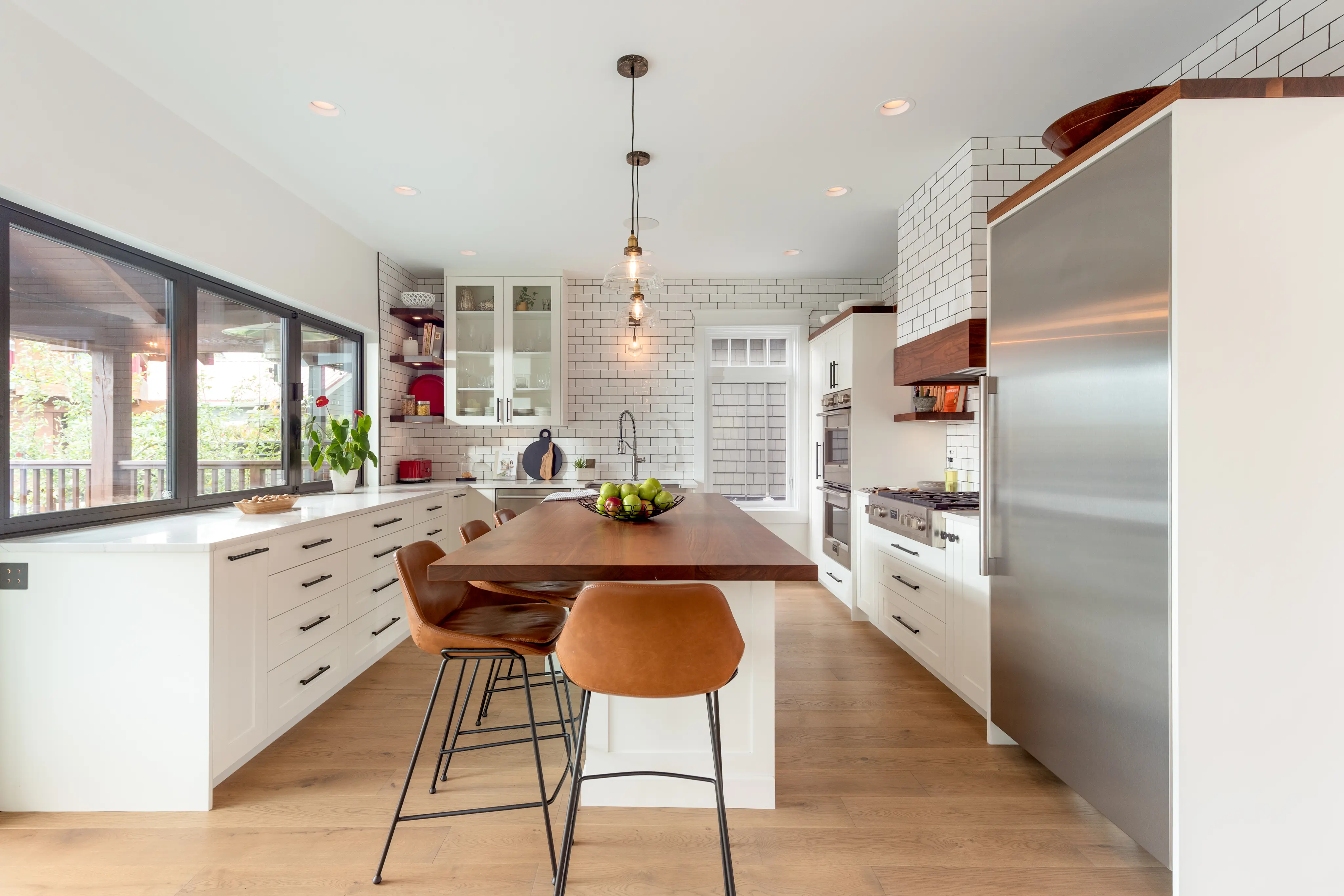 Bright modern kitchen with white cabinetry, wooden island countertop, brown leather bar stools, stainless steel refrigerator, and large window.