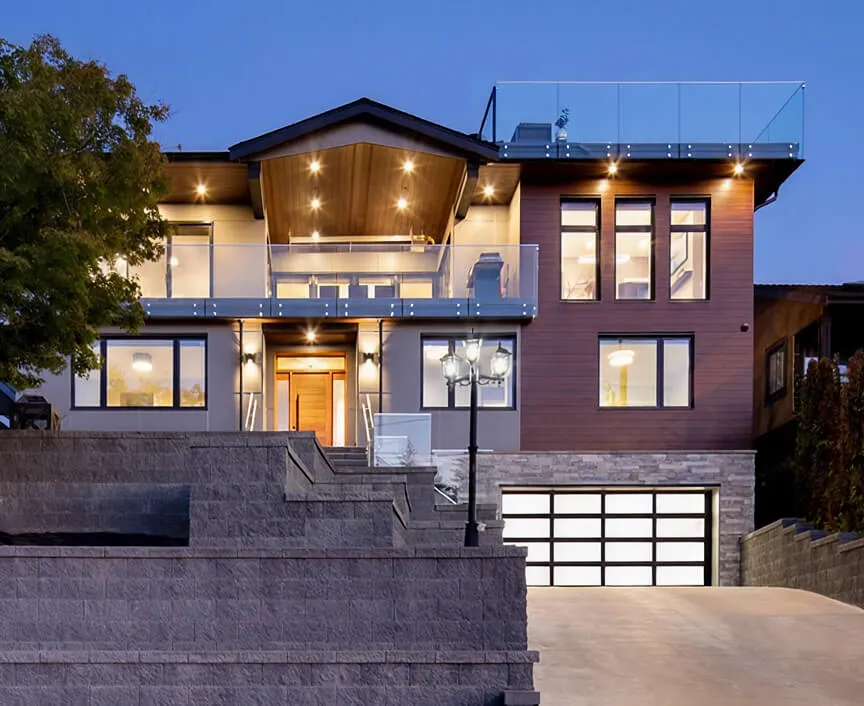 Modern two-story house with large windows, glass balcony, stone walls, and a driveway garage at dusk.