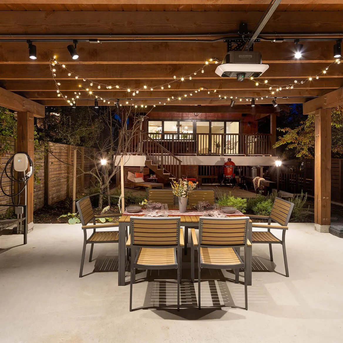 Outdoor dining area under wooden pergola with string lights, a table set for six, and a house with lit windows in the background at night.