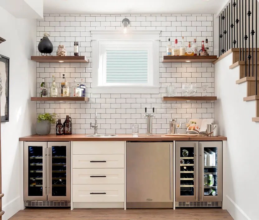 Modern home bar with white subway tiled wall, wooden countertop, stainless steel mini-fridges, sink, and liquor bottles on floating shelves.