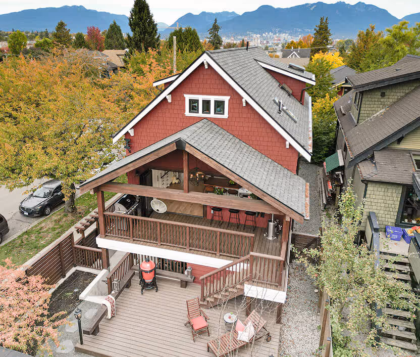 A red two-story house with a covered wooden balcony, outdoor seating area on the lower deck, surrounded by trees with mountains and a city skyline in the background.