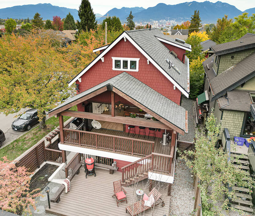 A red two-story house with a covered wooden balcony, outdoor seating area on the lower deck, surrounded by trees with mountains and a city skyline in the background.