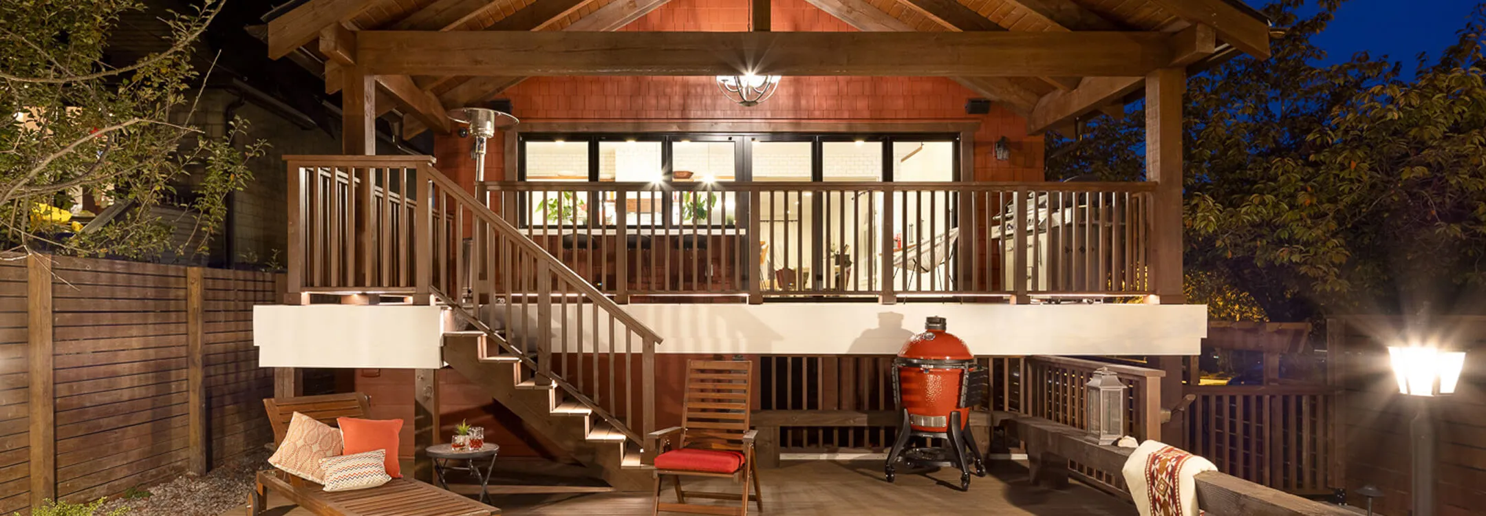 Evening view of a wooden backyard deck with stairs, seating, a red grill, and illuminated indoor area behind glass doors.