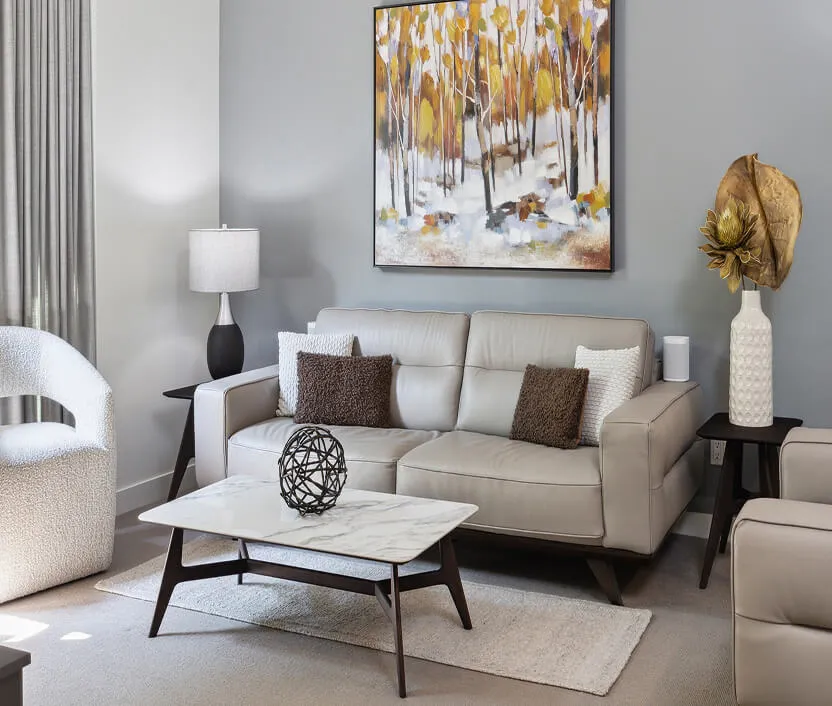 Modern living room with beige leather sofa adorned with brown and white cushions, a white armchair, marble coffee table with decorative sculpture, side tables with lamp and vase, and abstract autumn forest painting on gray wall.