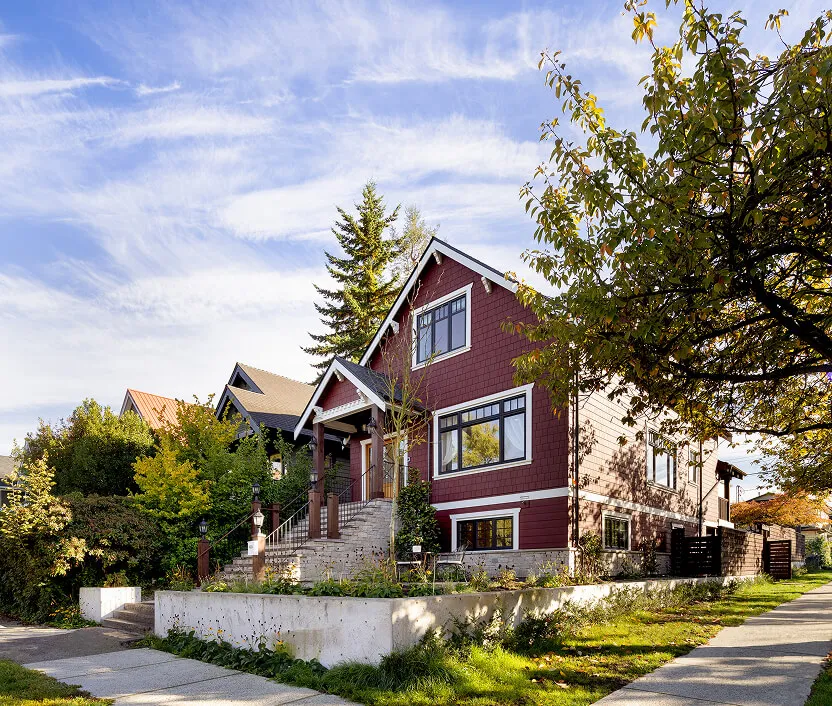 Two-story house with maroon siding, large windows, and a front porch with stairs, surrounded by trees and greenery on a sunny day.