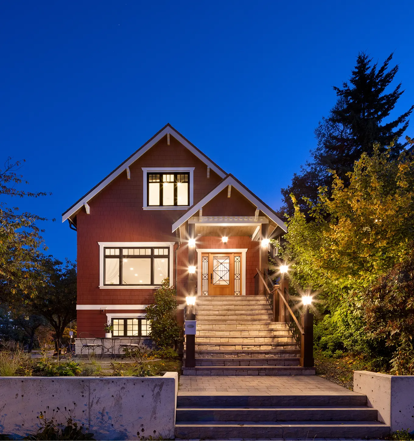 Red house with lit windows, front stairs, and surrounding trees at dusk under a clear blue sky.
