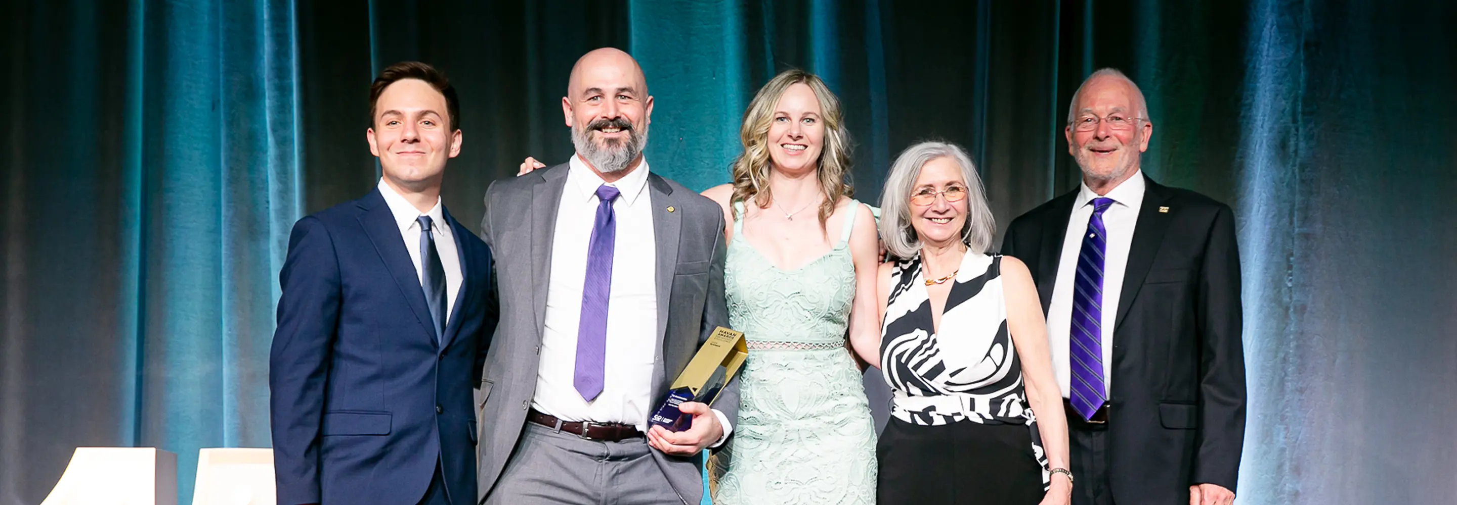 Five people standing in a row at an event, one holding a golden and blue award trophy, all dressed in formal attire and smiling.