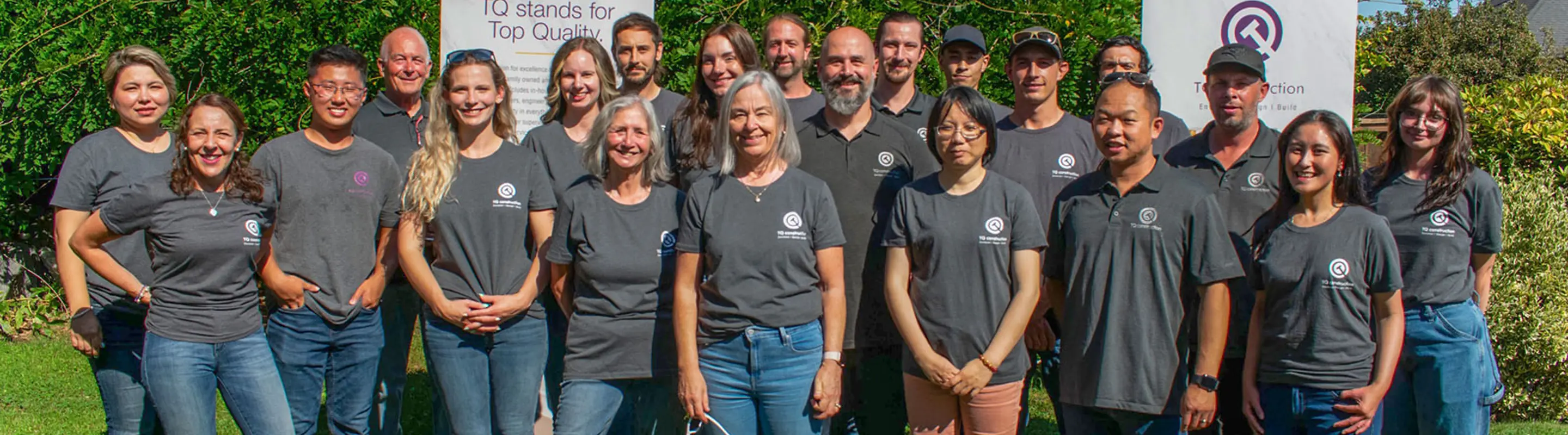 Group of diverse adults standing outdoors in front of greenery, all wearing matching gray company t-shirts with a logo.