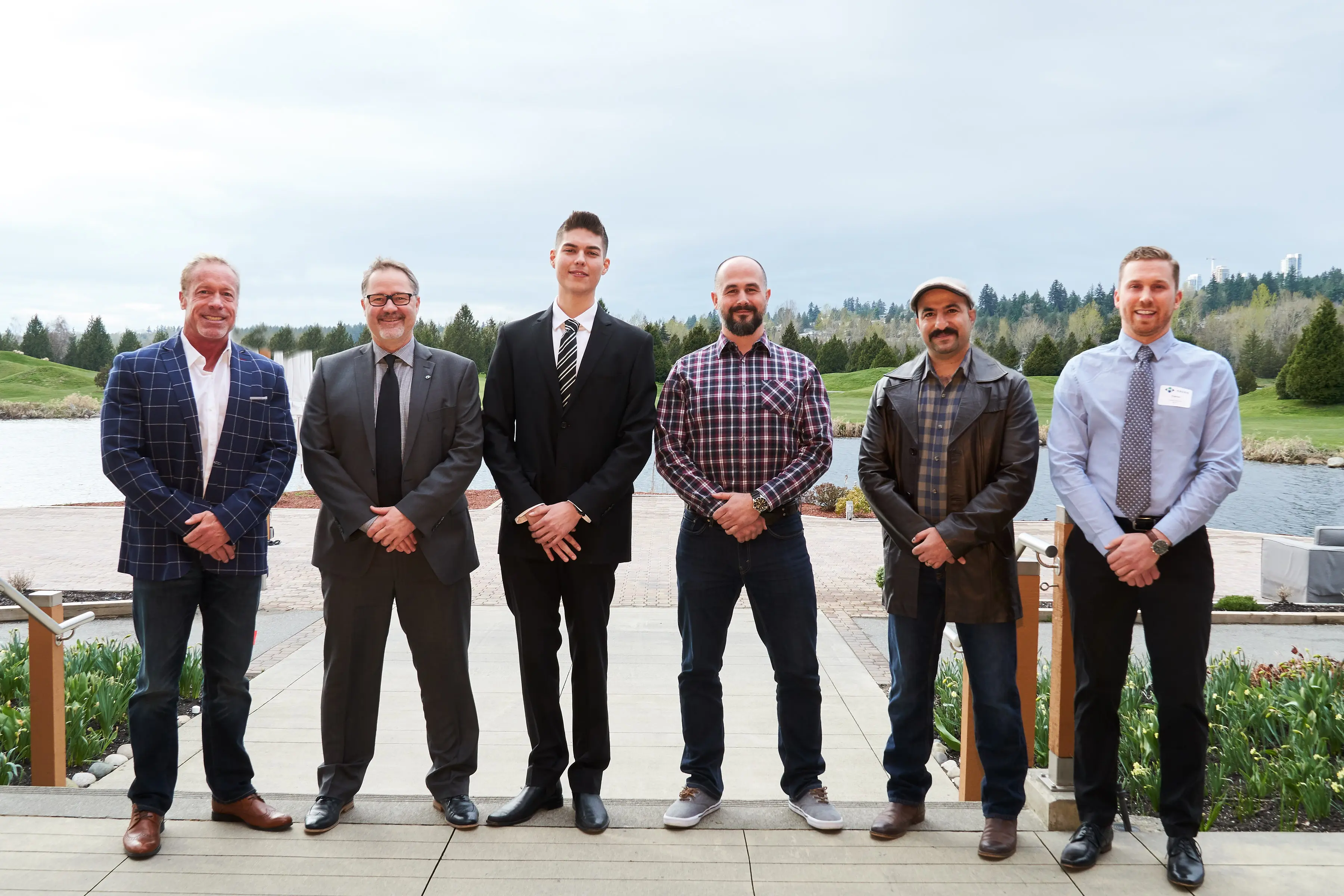 Six men standing side by side outdoors in front of a pond and greenery, dressed in a mix of business and casual attire.