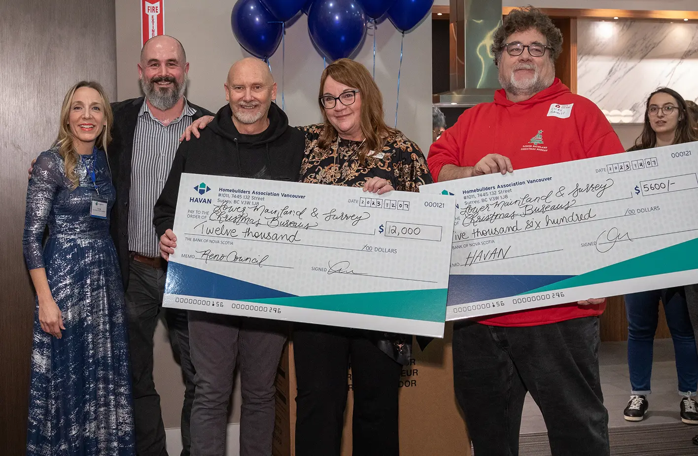 Five people standing indoors holding two large ceremonial checks for donation amounts of $12,000 and $5,600 made out to Lower Mainland & Surrey Christmas Bureaus.