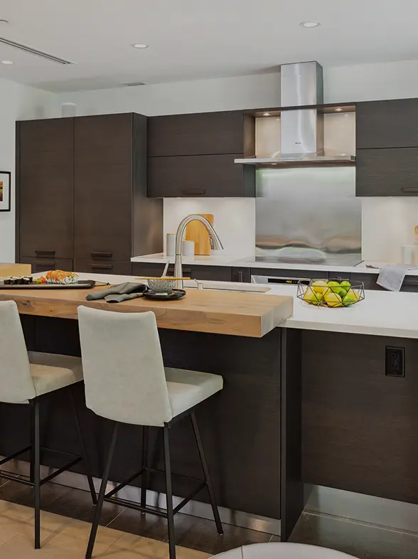 Modern kitchen with dark cabinets, a wooden breakfast bar with two beige stools, a stainless steel range hood, and a bowl of green apples on the counter.