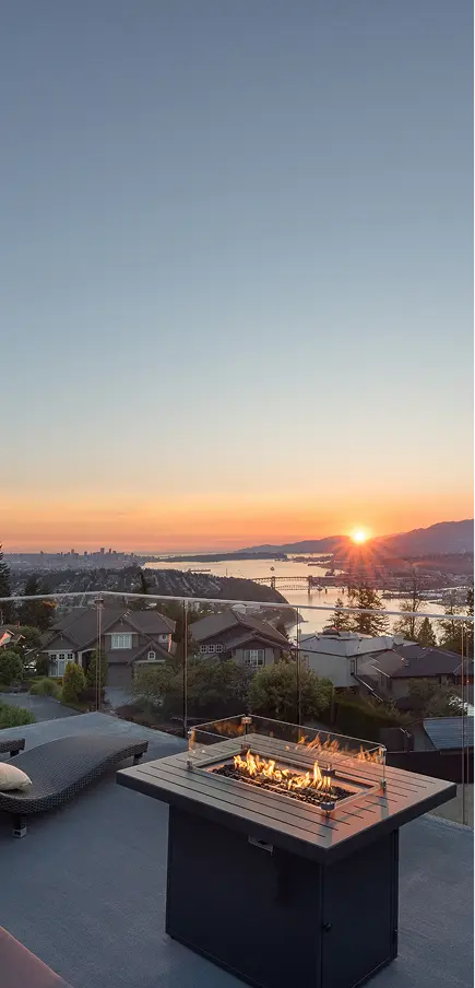 Modern outdoor patio with fire pit and lounge chair overlooking houses, a river, and mountains at sunset.