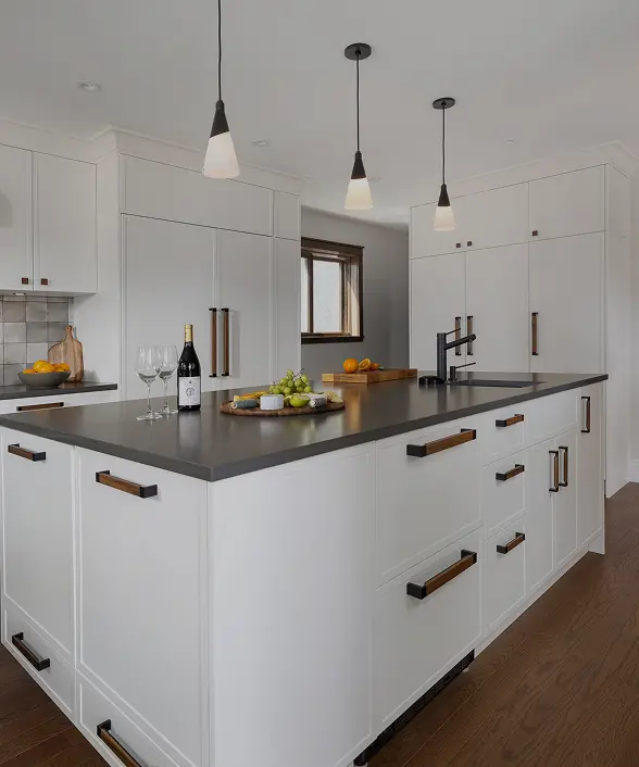 Modern kitchen with white cabinetry, a large island with dark countertop, three pendant lights, and a bottle of wine with two glasses on the island.