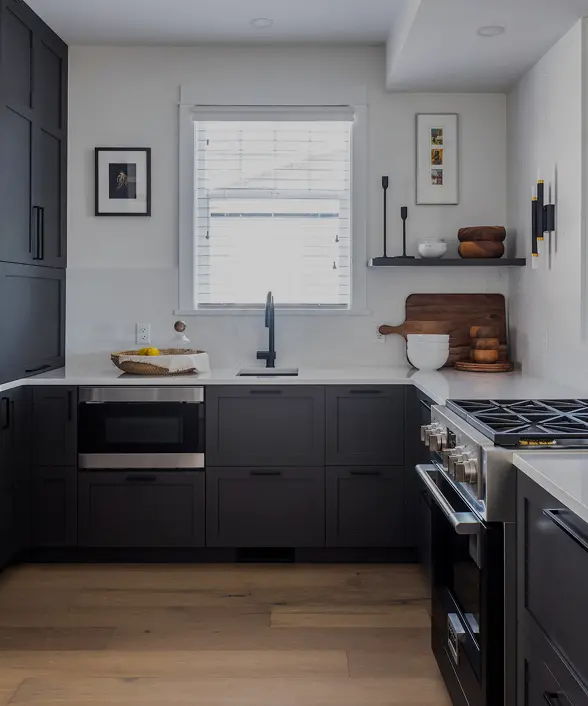 Modern kitchen with black cabinets, stainless steel oven, microwave, a window with white blinds, and wooden floor.