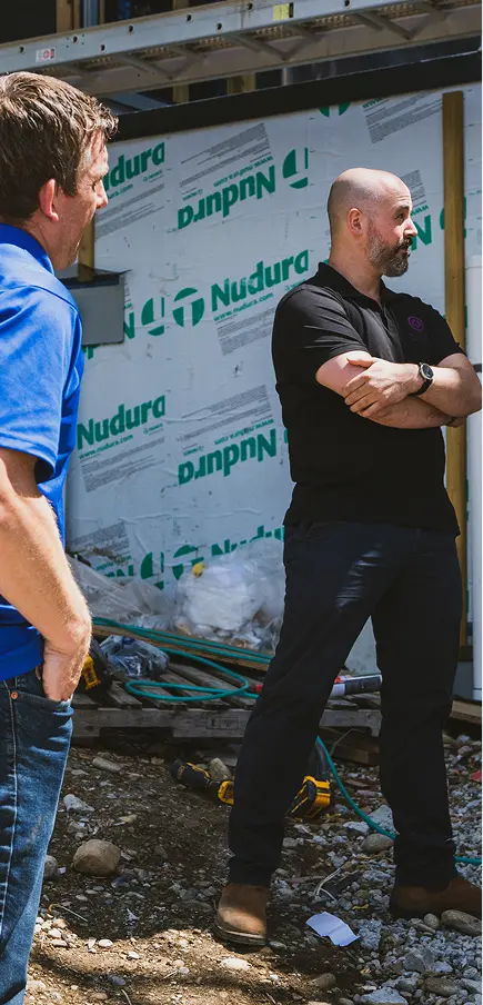 Two men talking at a construction site with building materials and exposed framework behind them.