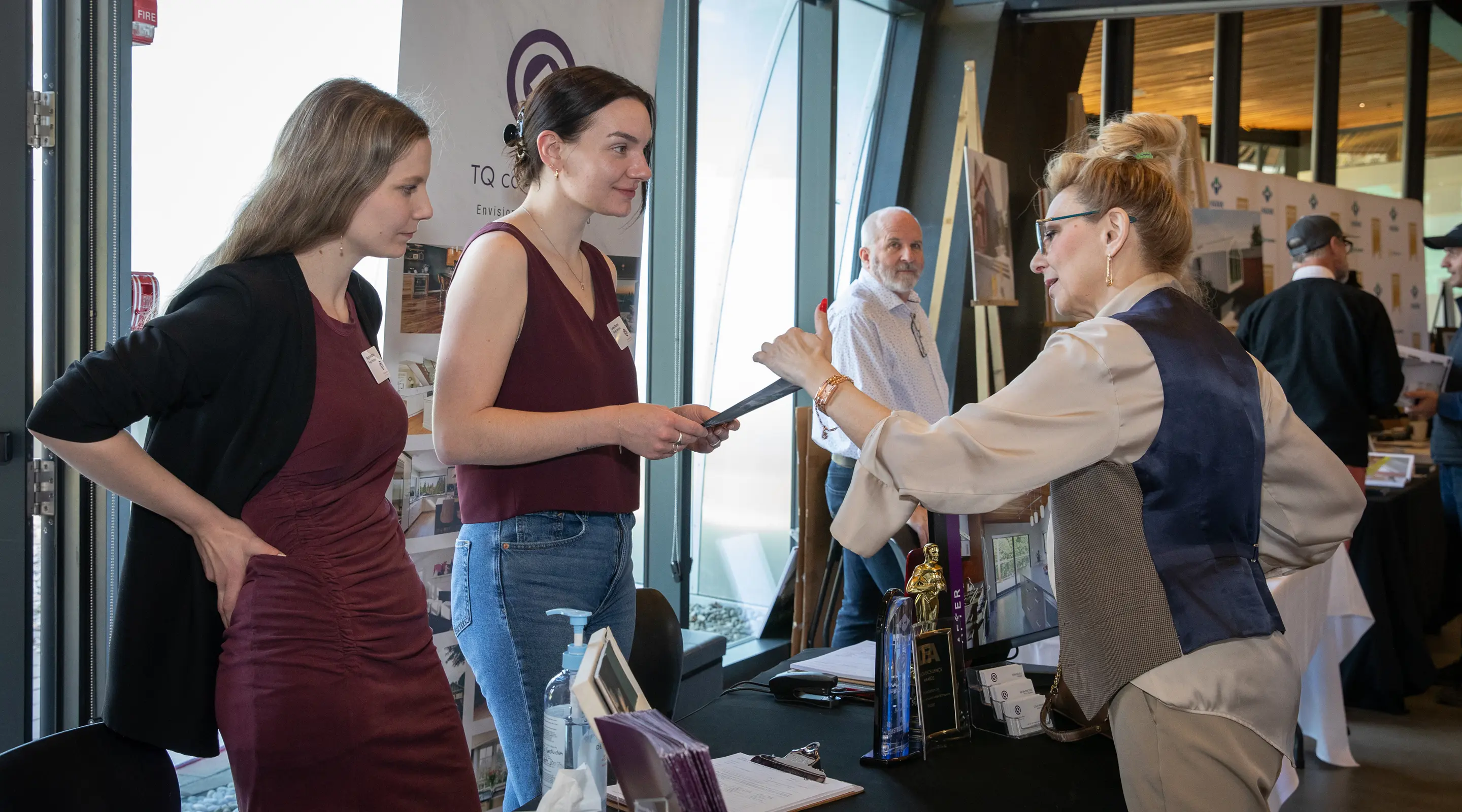 Three women and a man engaged in conversation at a booth with brochures and awards in a well-lit indoor event space.