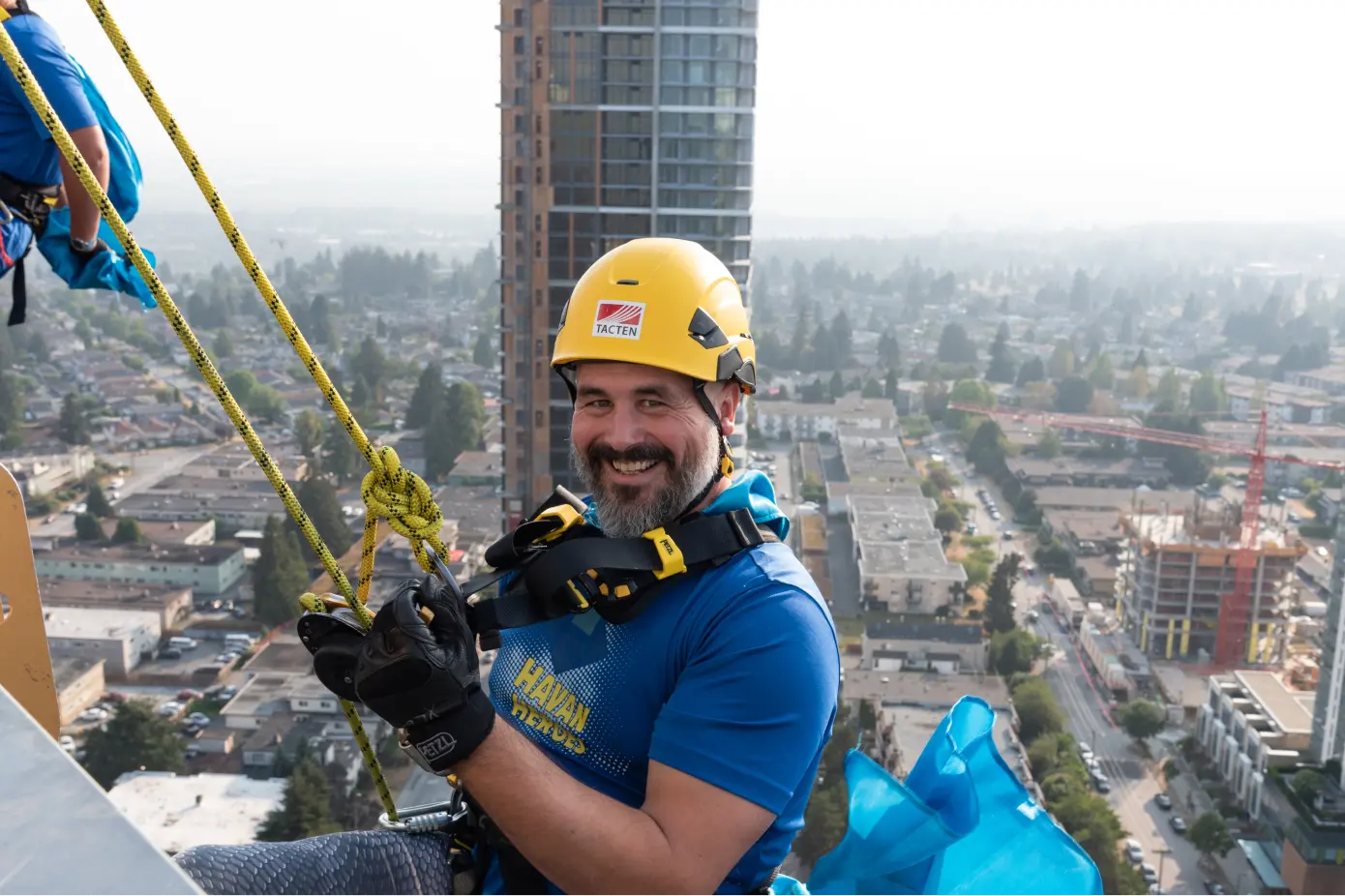 Smiling man wearing a yellow safety helmet and blue shirt rappel down a tall building overlooking an urban cityscape.