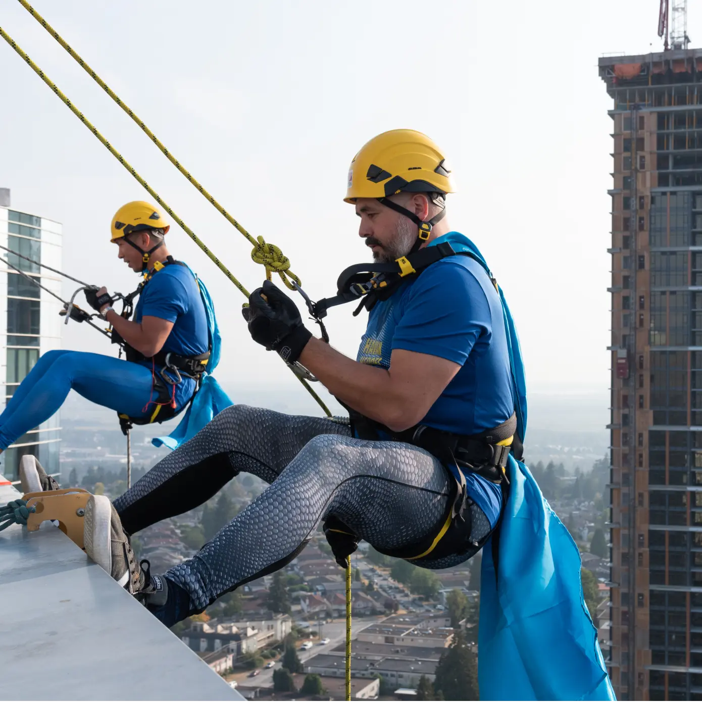 Two workers in yellow helmets and blue shirts rappelling down a building with safety harnesses and ropes.