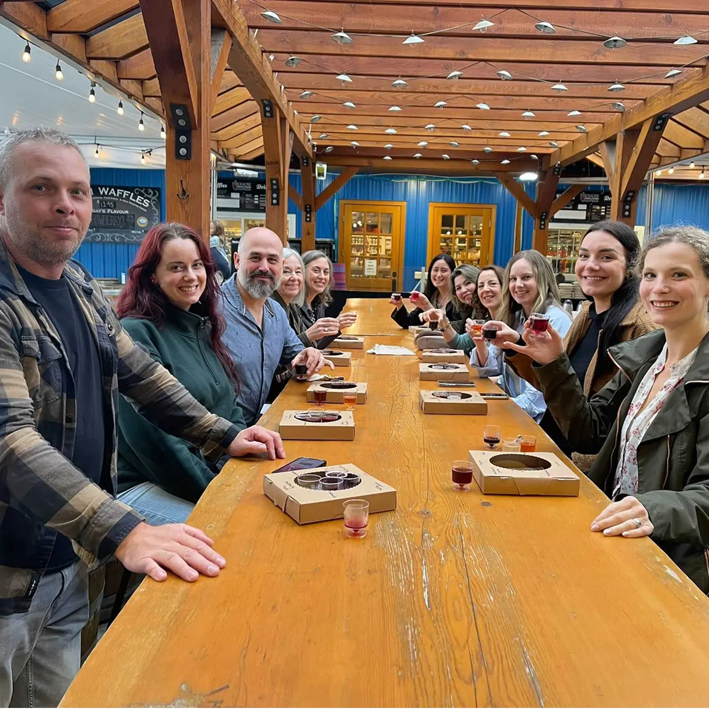 Group of people sitting at a long wooden table in a covered outdoor area, holding small glasses with drinks and smiling at the camera.