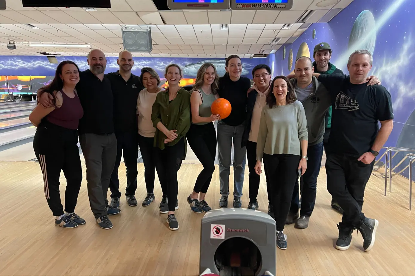 Group of twelve people smiling and posing together on a bowling alley with a space-themed mural on the wall.