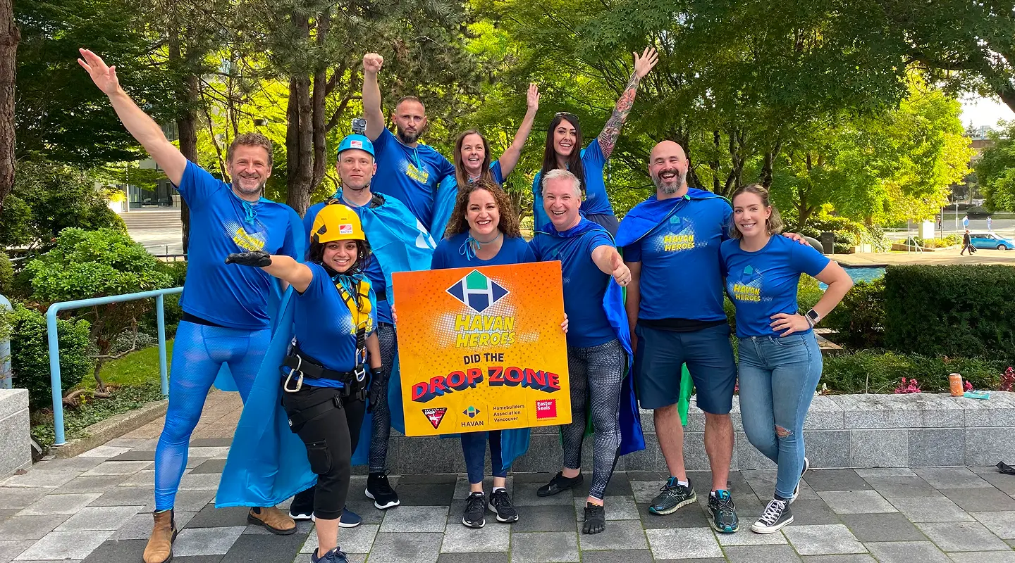 Group of ten people outdoors wearing blue Havan Heroes shirts and capes, smiling and holding a sign that says 'Havan Heroes did the Drop Zone.'