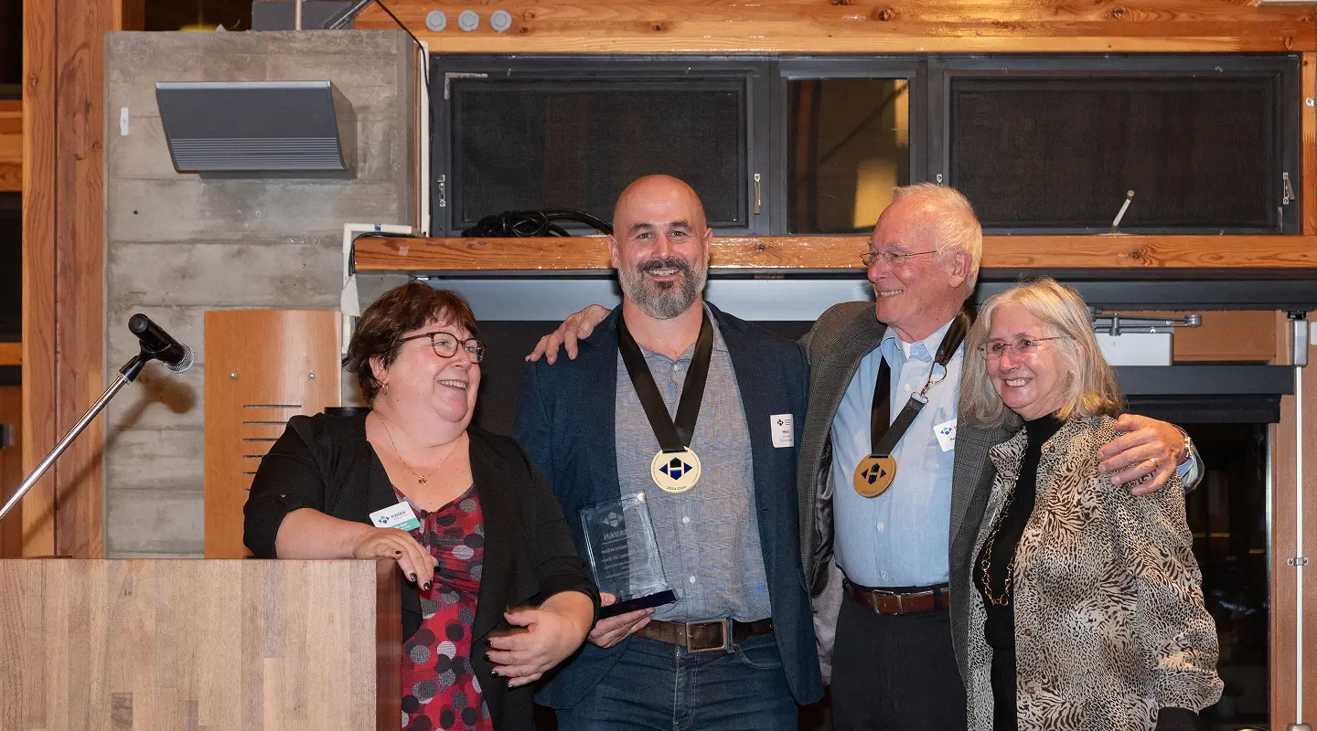 Four people smiling at an indoor event; two men in the center wear medals, one holds a glass award, and two women stand on either side.