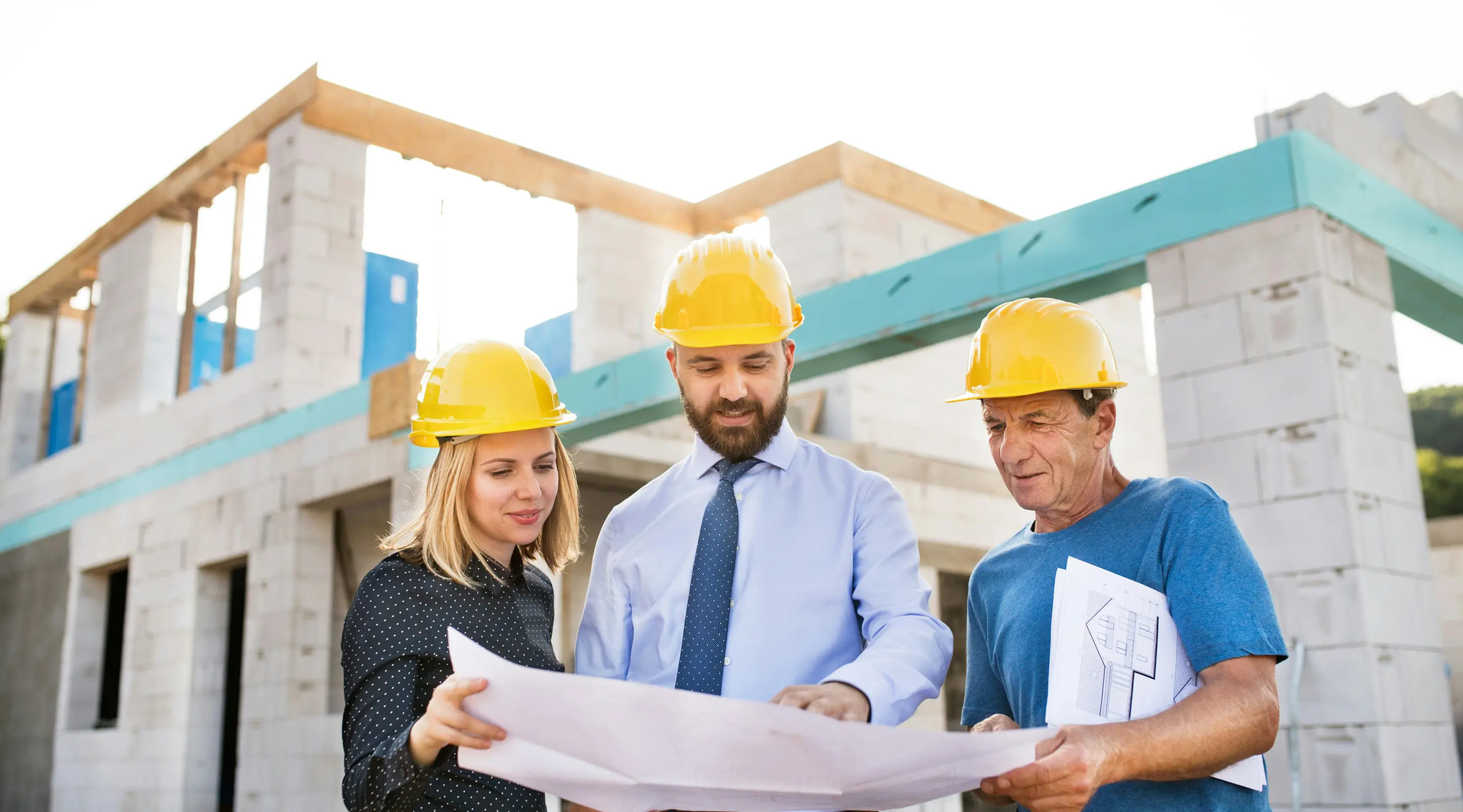 Three construction professionals wearing yellow hard hats reviewing building plans in front of a partially constructed building.