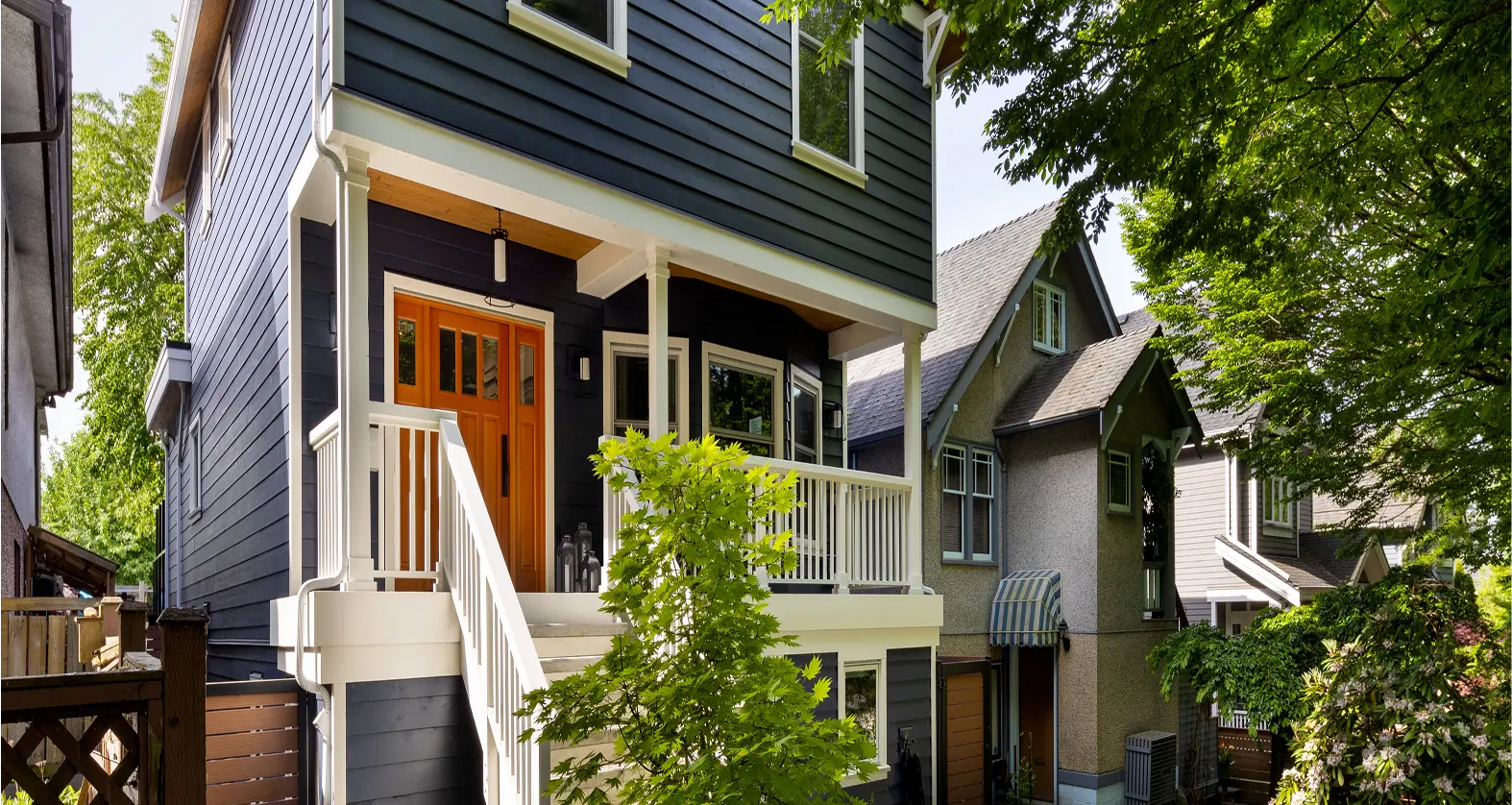 Modern dark blue house with white trim, orange front door, and white staircase, surrounded by lush green trees and neighboring houses.