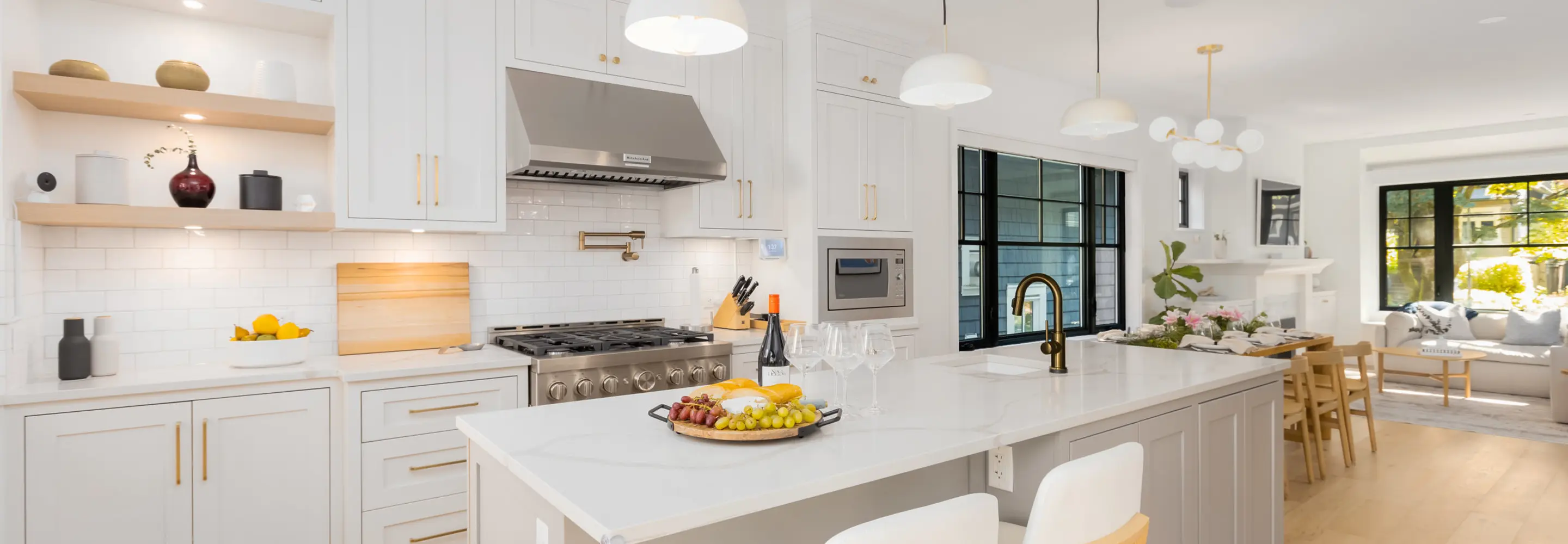Bright modern kitchen with white cabinetry, marble countertop island with fruit and wine, stainless steel stove, and black-framed windows overlooking a living area.