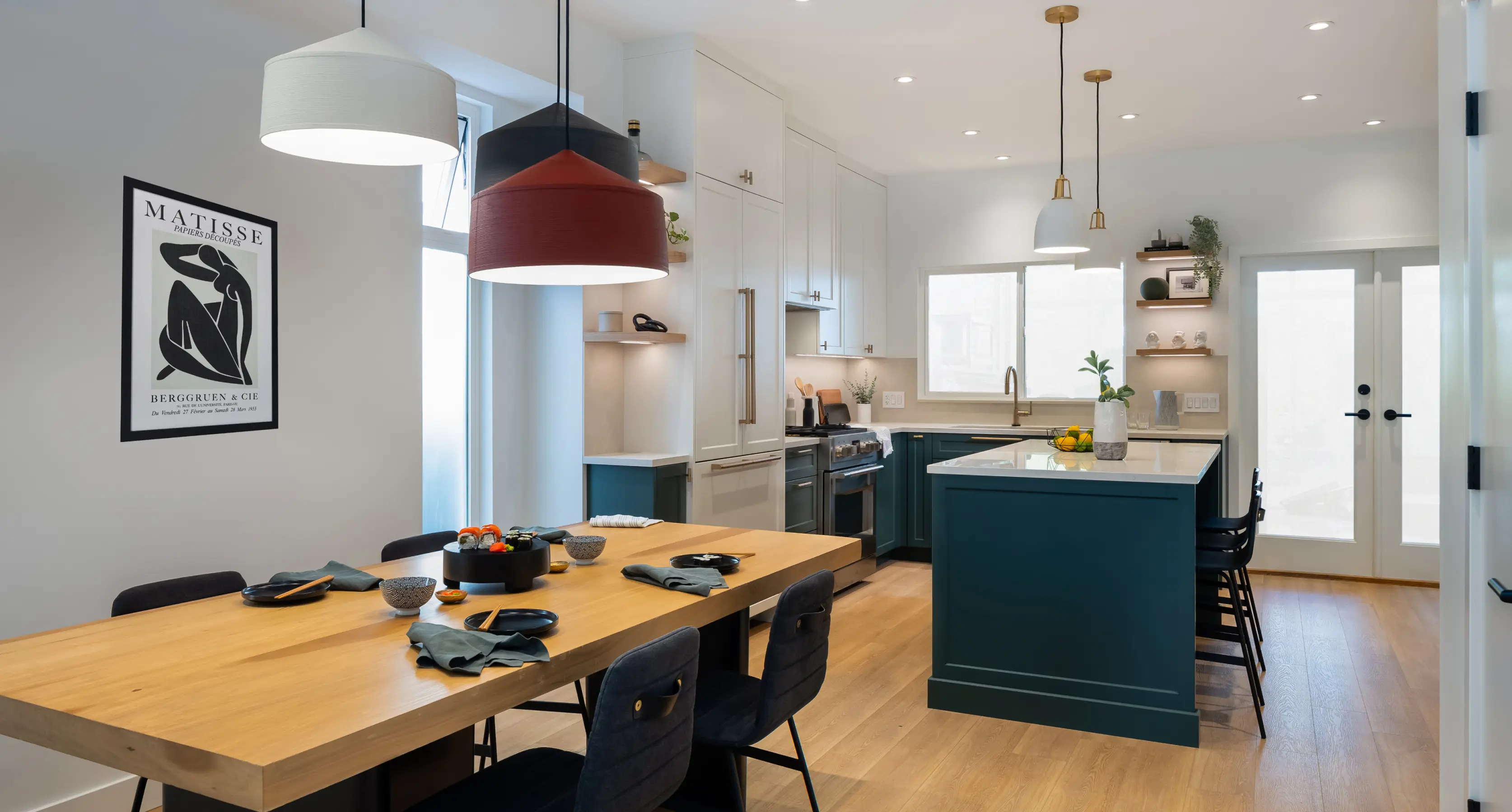 Modern kitchen and dining area with wooden table set for sushi, teal blue cabinets, white countertops, and three hanging pendant lights.