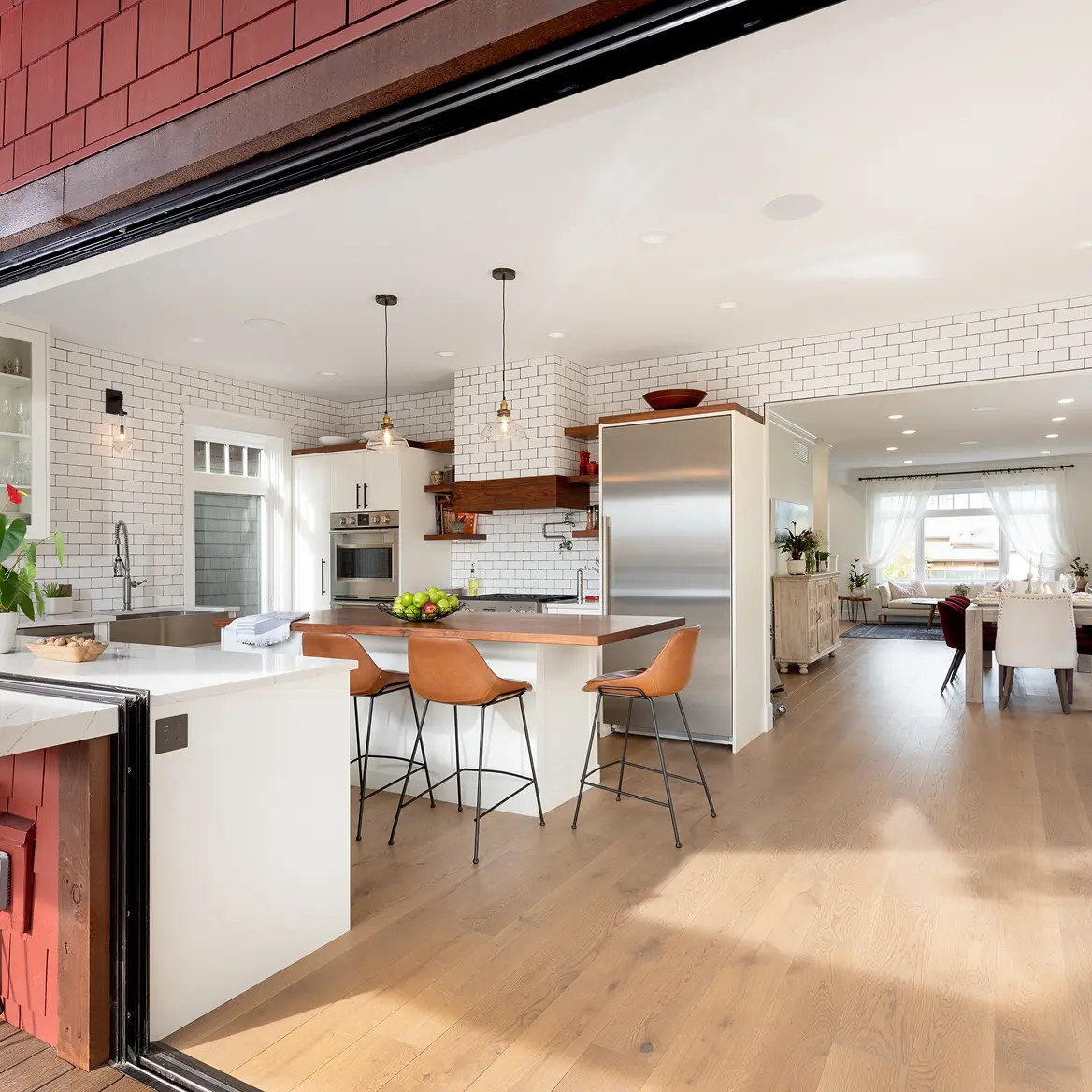 Bright modern kitchen with white subway tile walls, wood floor, leather bar stools, and a stainless steel refrigerator opening to a dining and living area with large windows.