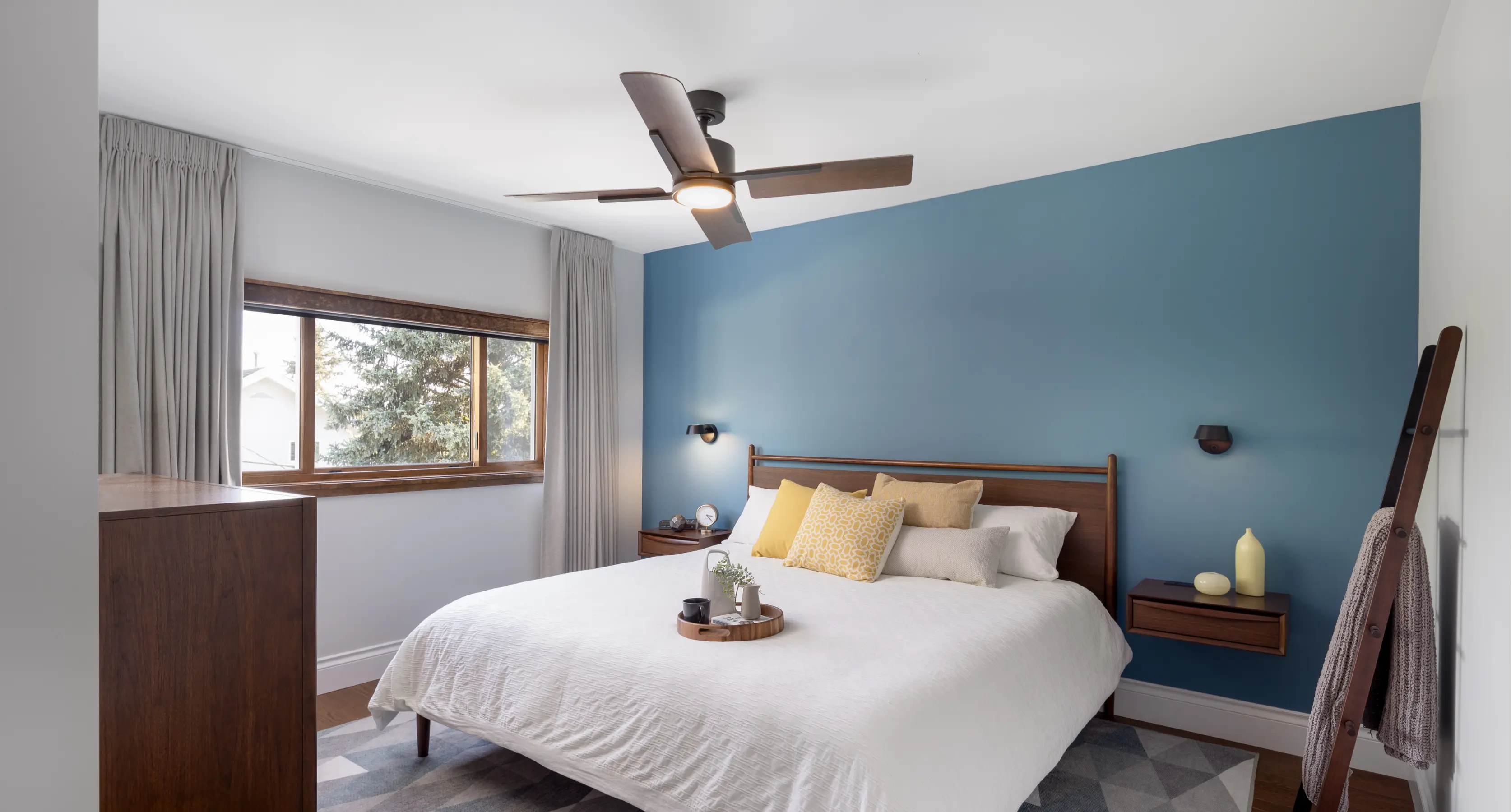 Modern bedroom with a wooden bedframe, white bedding, yellow and beige pillows, blue accent wall, ceiling fan, and wooden furniture.