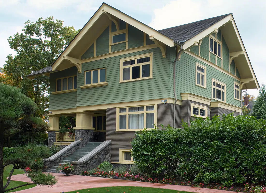 Two-story craftsman-style house with green and brown siding, stone stairs, and lush landscaping.
