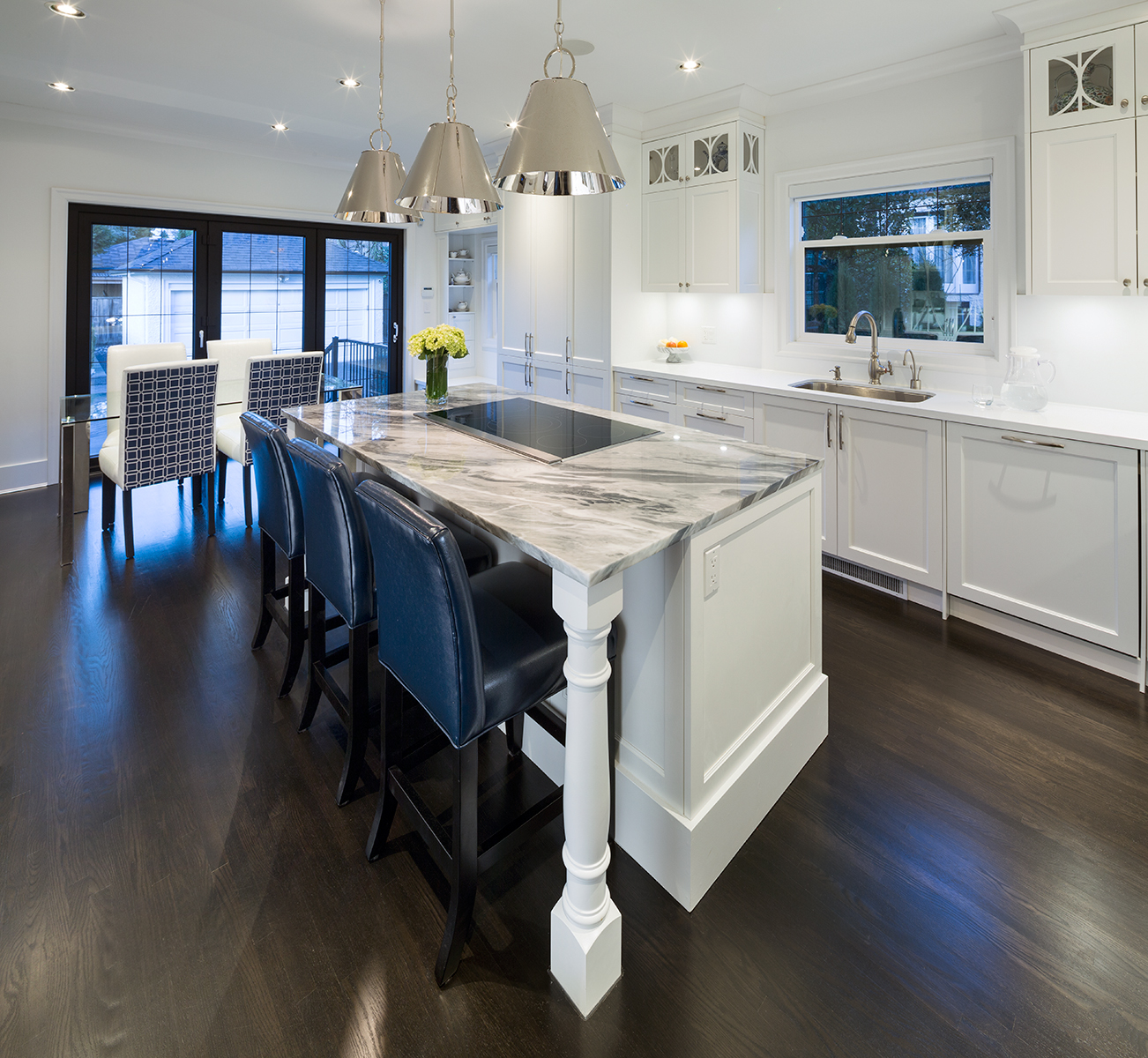 Modern kitchen with marble island countertop, dark wood floor, white cabinets, and blue and white chairs.