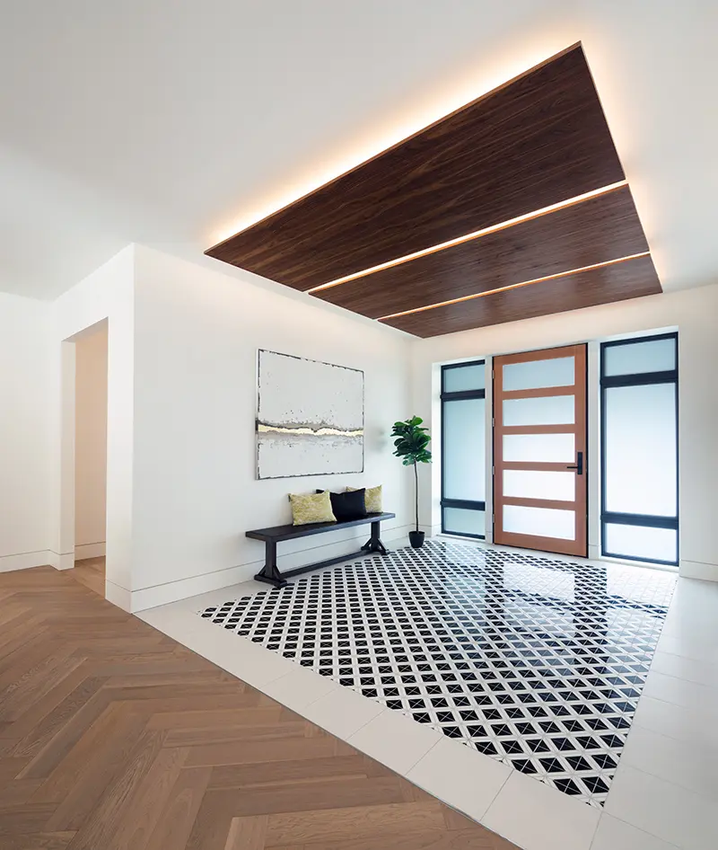 Modern entryway with wooden front door, geometric black-and-white tiled floor area, wood-paneled ceiling with lighting, bench with cushions, and a potted plant.