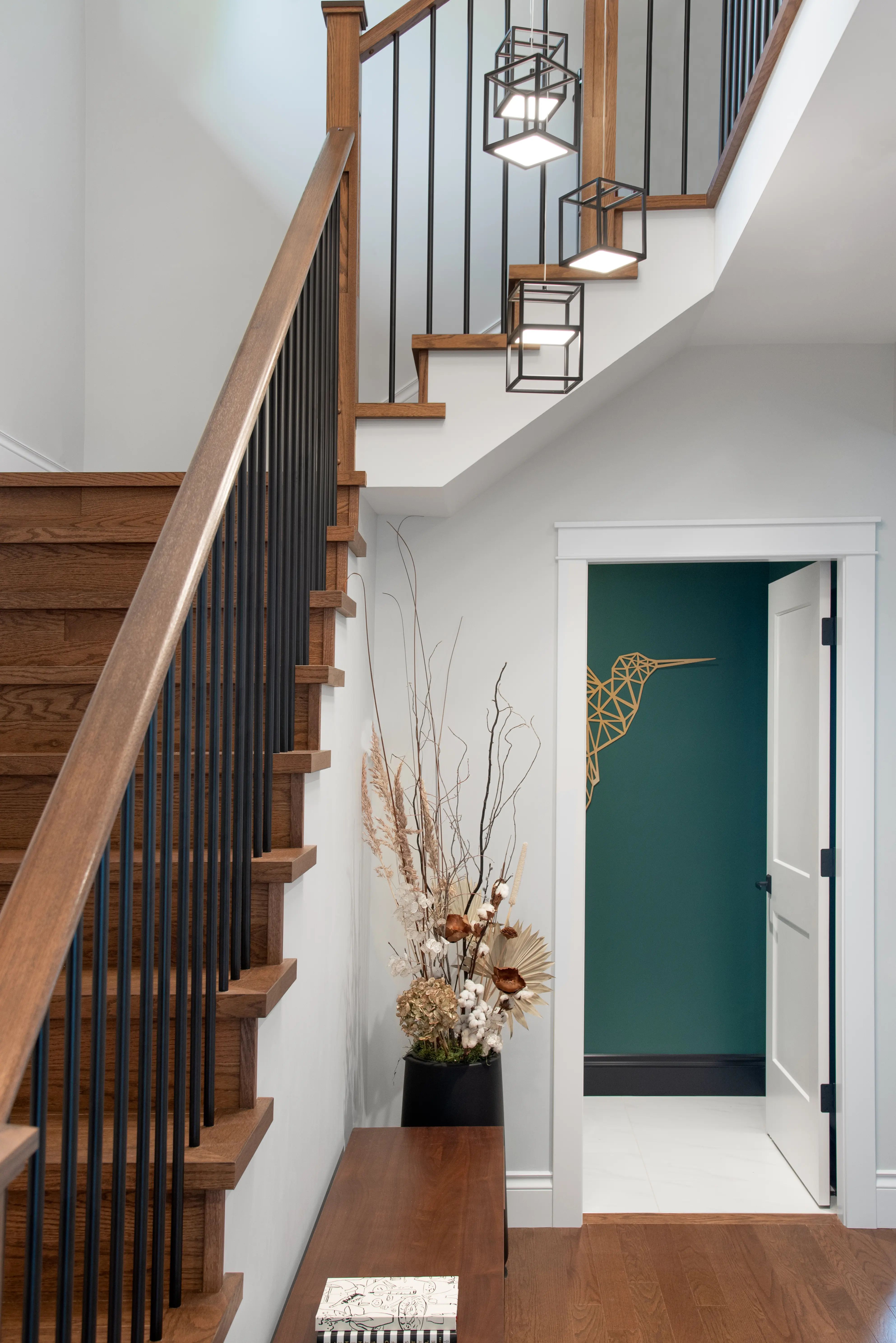 Modern wooden staircase with black metal balusters next to a bench, a vase with dried flowers, and a partially open door revealing a wall with a geometric bird design.