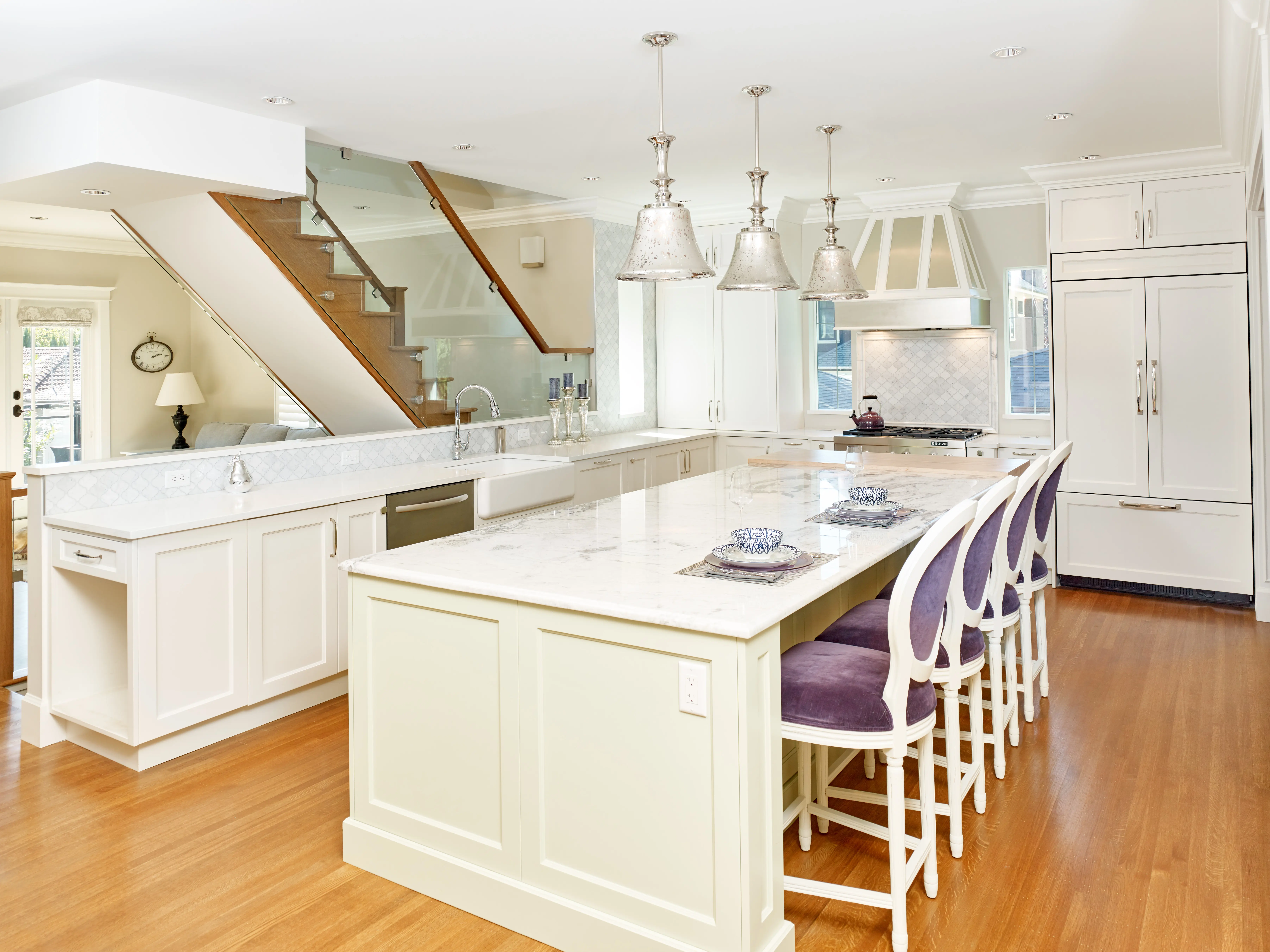 Bright modern kitchen with white cabinetry, large marble island, purple cushioned chairs, wood flooring, and three pendant lights.