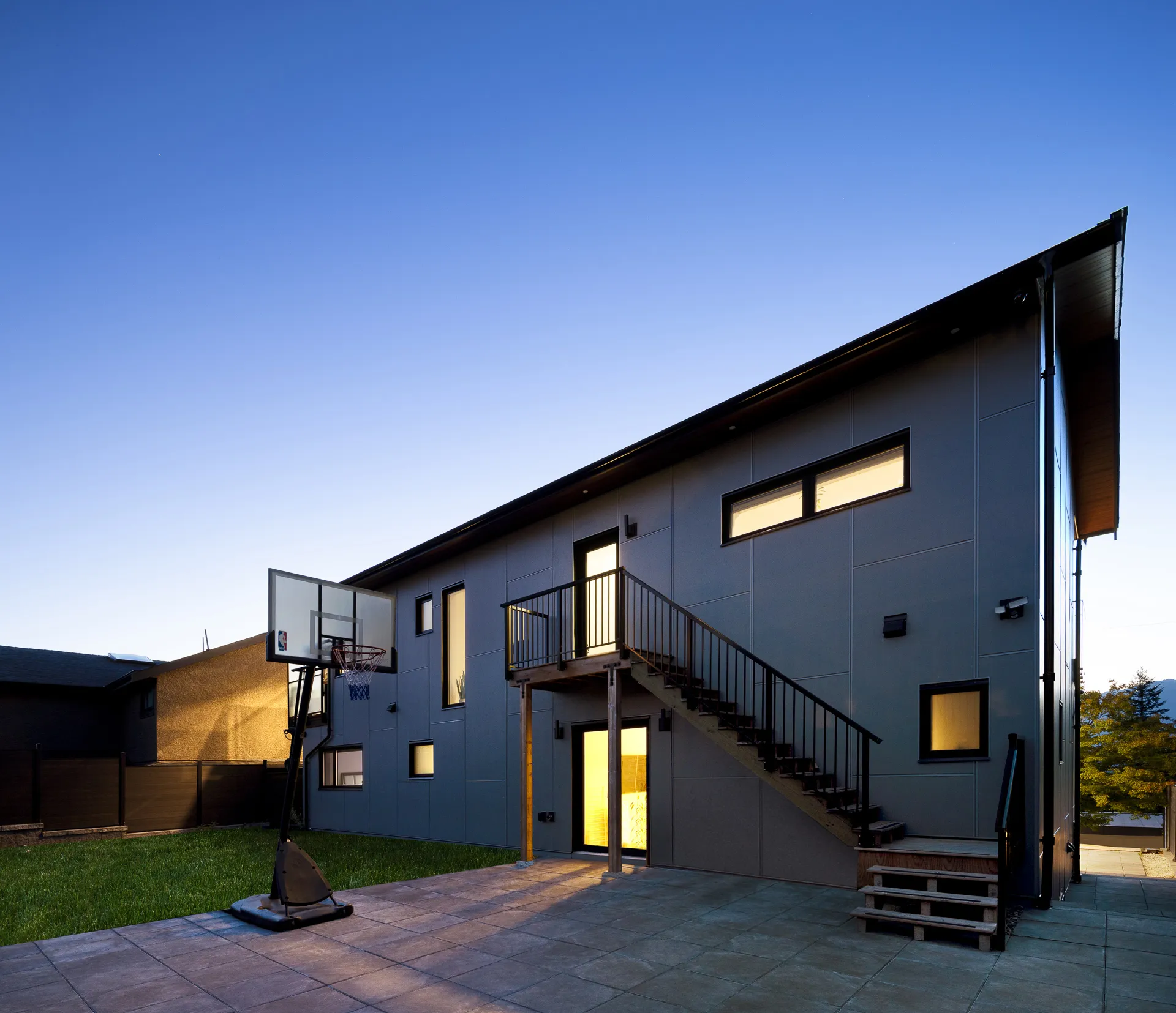 Modern two-story gray house with outdoor staircase, illuminated doors and windows, and a basketball hoop on paved patio at dusk.