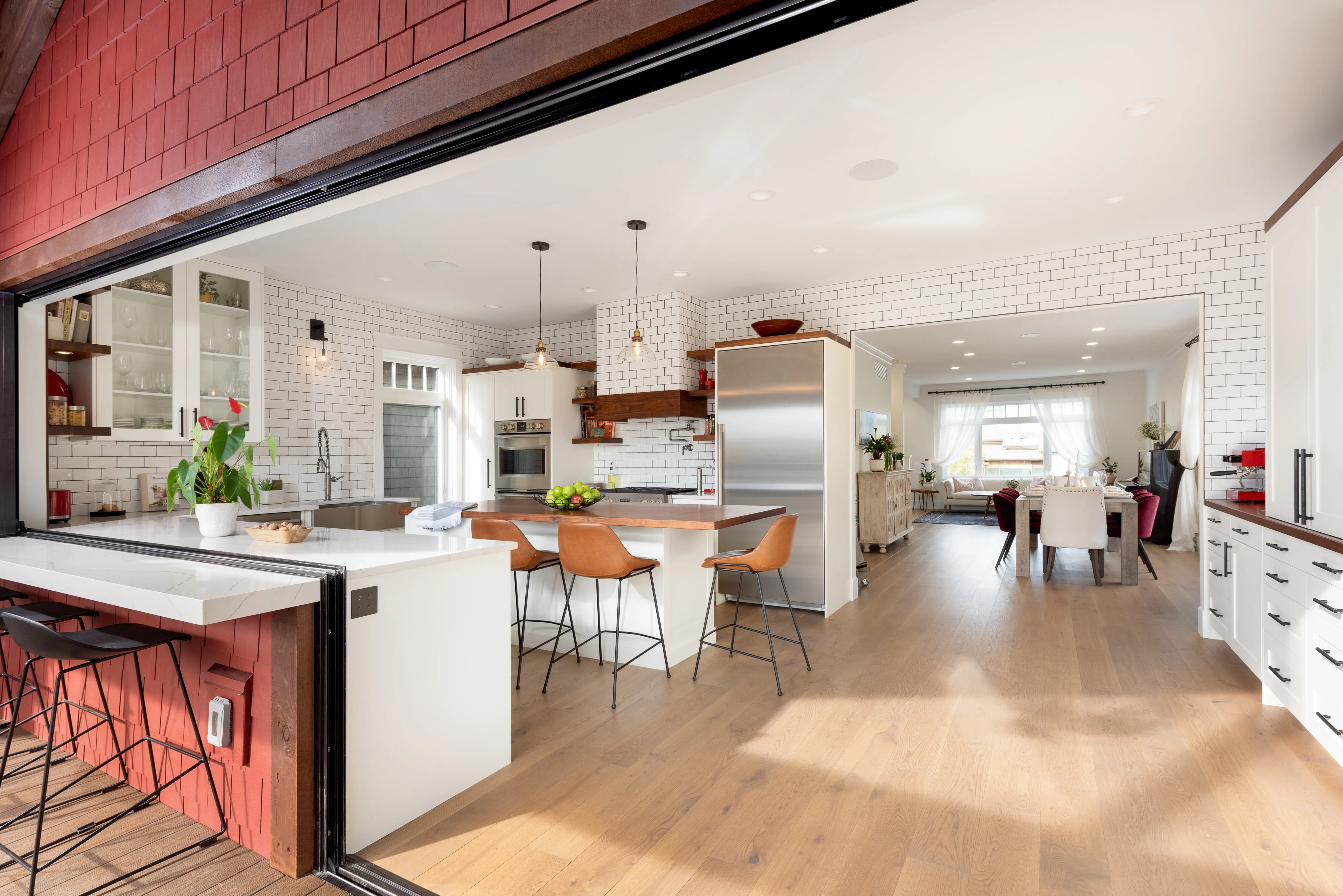 Modern open kitchen with white subway tile walls, wooden floor, a large island with brown leather bar stools, and an adjoining dining area with red and white chairs.