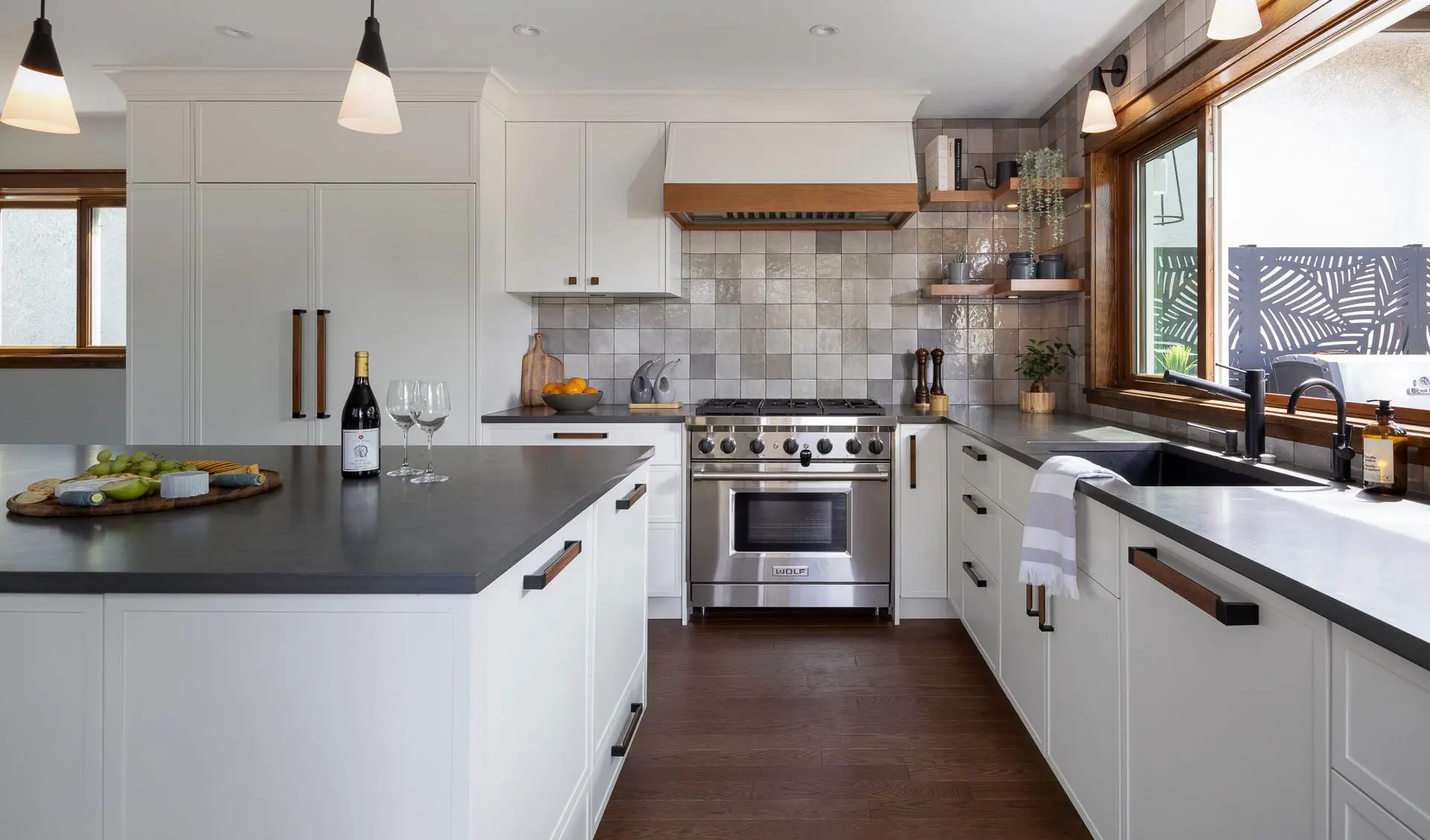 Modern kitchen with white cabinetry, dark countertops, stainless steel stove, a wine bottle with two glasses on island, and a window over the sink.