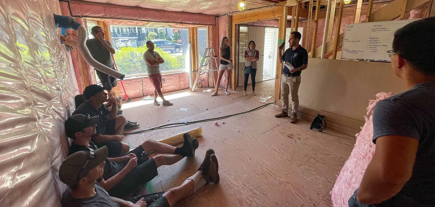 Group of people in casual clothing inside unfinished house during a meeting or briefing, with exposed insulation and wooden floor.