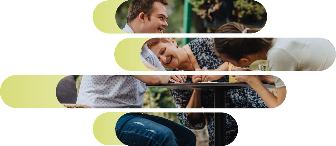 Four people sitting outdoors at a table, smiling and leaning in to look at something on the table together.