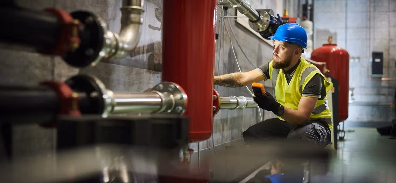 A professional technician wearing a hard hat and high-visibility vest kneels in a large boiler room, holding a meter to check pipes and industrial heating equipment.