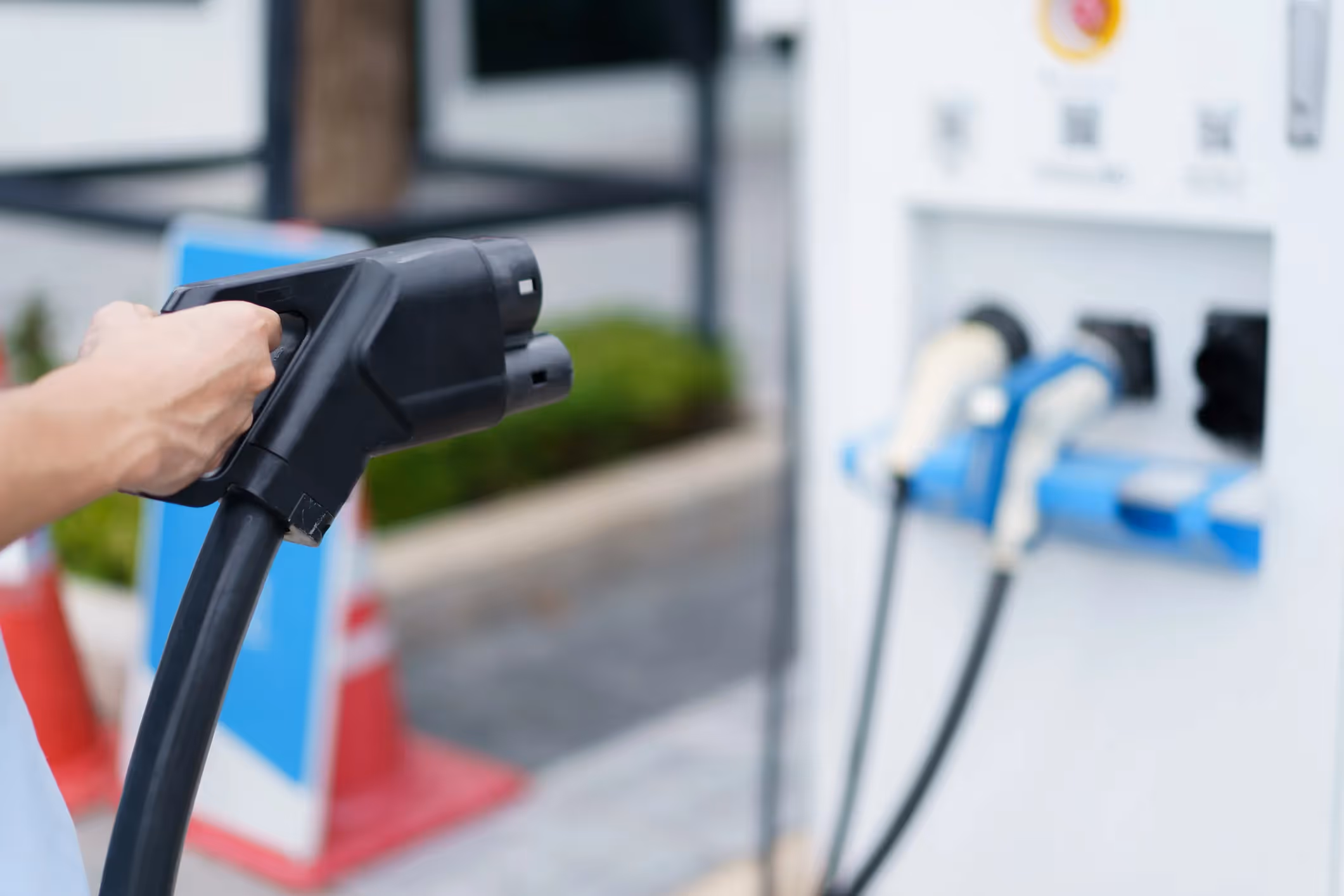 Person's hand holding a dual-port black EV charging connector near a public charging pedestal.