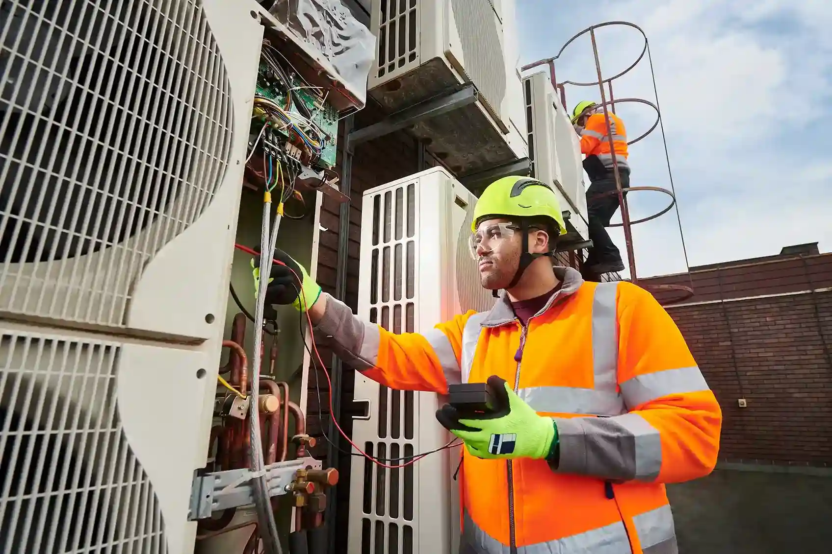 An electrician in a high-visibility jacket and helmet checks wiring on a large outdoor air conditioning unit with a multimeter.
