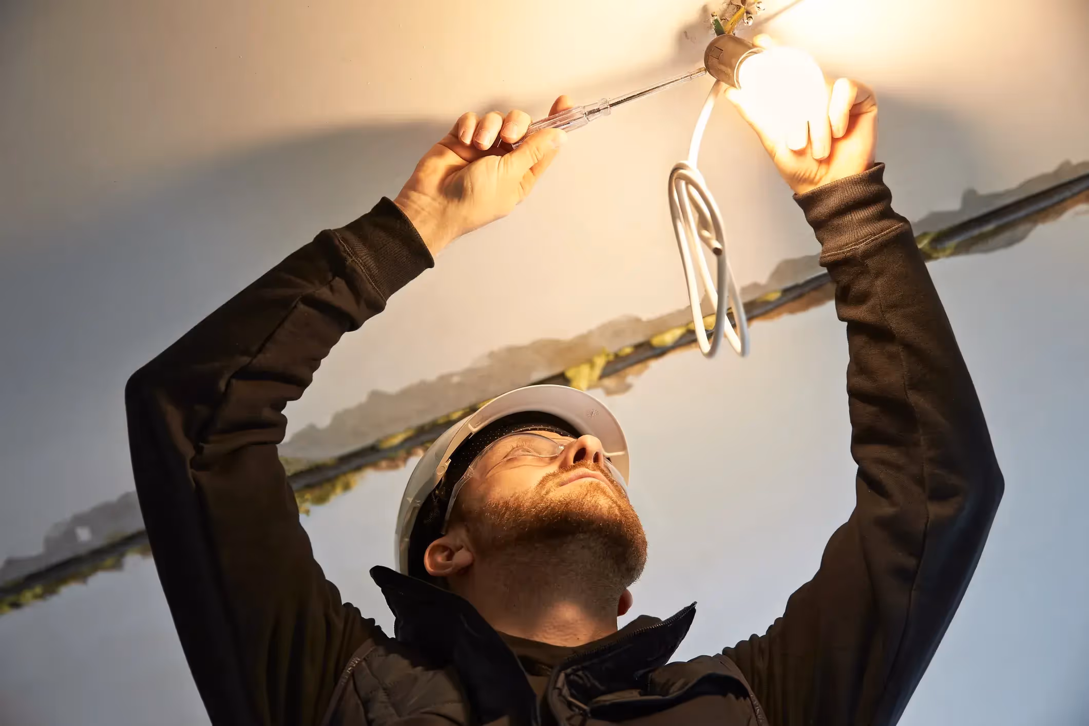 A low-angle shot of an electrician in a hard hat and safety glasses holding a glowing light bulb and a tool to work on a ceiling light fixture.